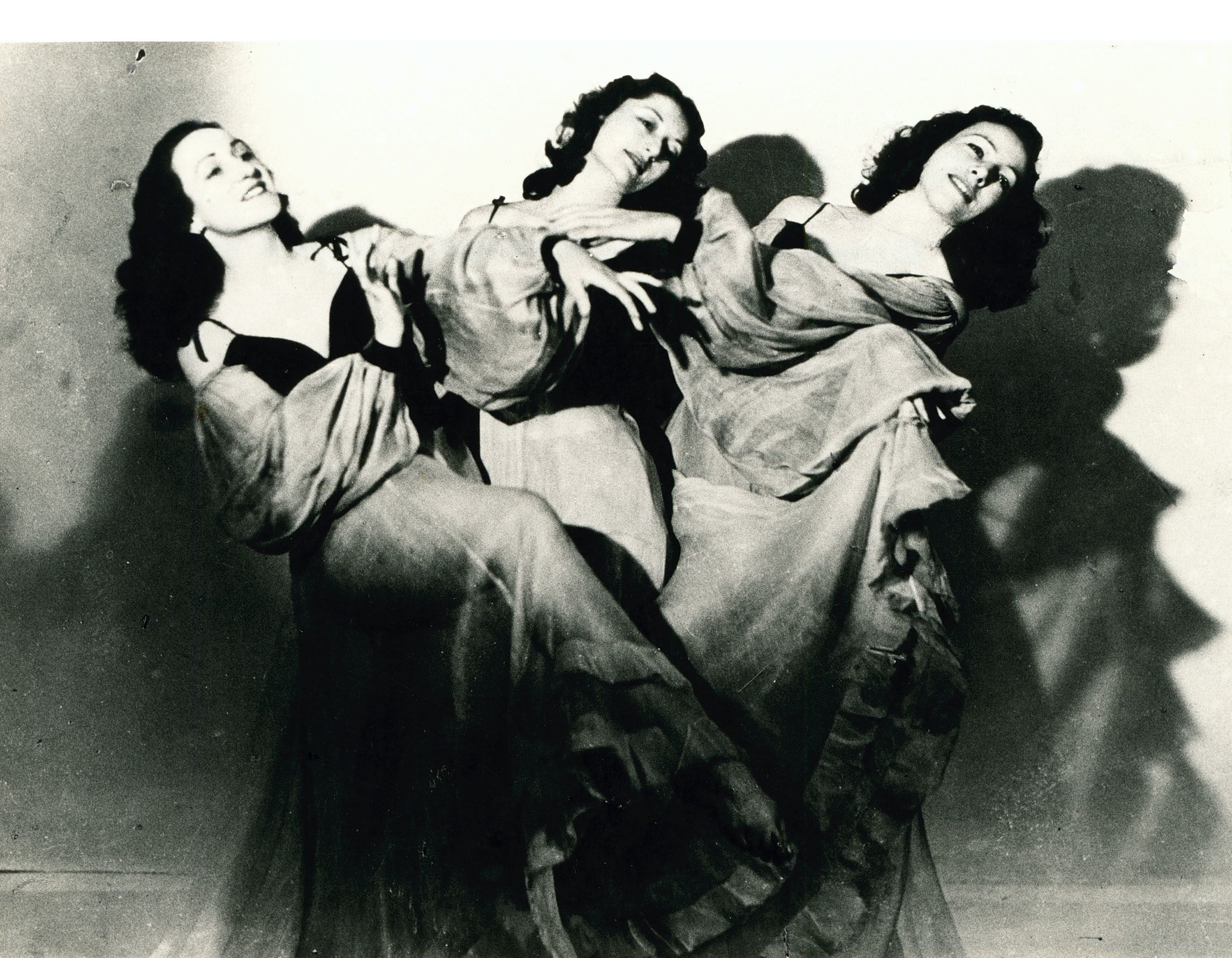 Black and white mid-shot image of three female dancers, wearing draped fabric from the shoulders downwards. One dancer is leaning towards the left and two towards the right side.