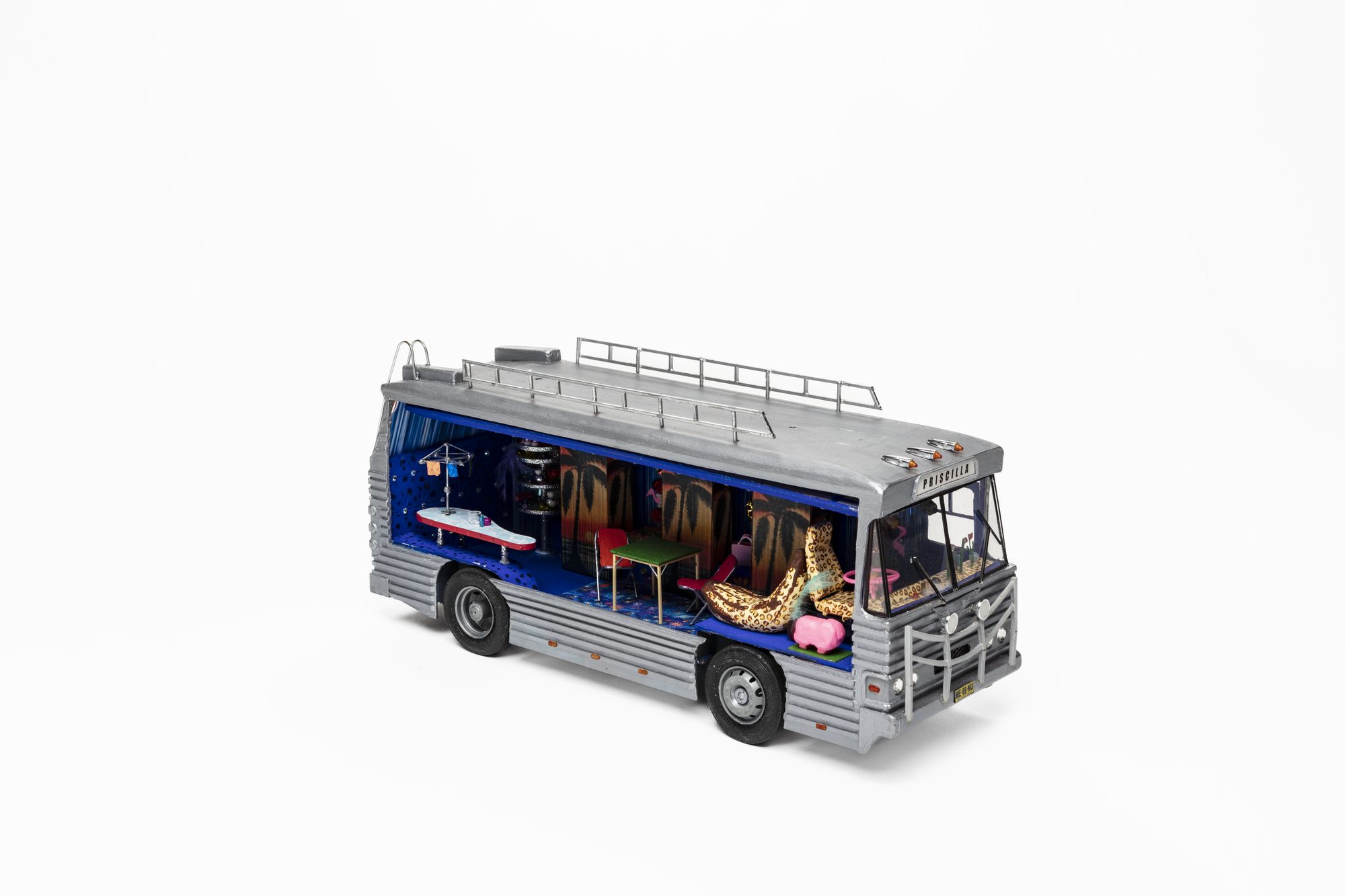 Three quarter side view of silver-coloured model bus with open panel to interior. Inside features brightly coloured miniature furnishings including of a rounded edge bench, hills hoist clothesline, table and two chairs, leopard print covered driver’s seat and banana-shaped lounge, pink sheep and steering wheel. Three panels with an image of a sunset and palm tree hanging on long interior wall of bus. A sign above the windscreen reads ‘Priscilla’ in black lettering.