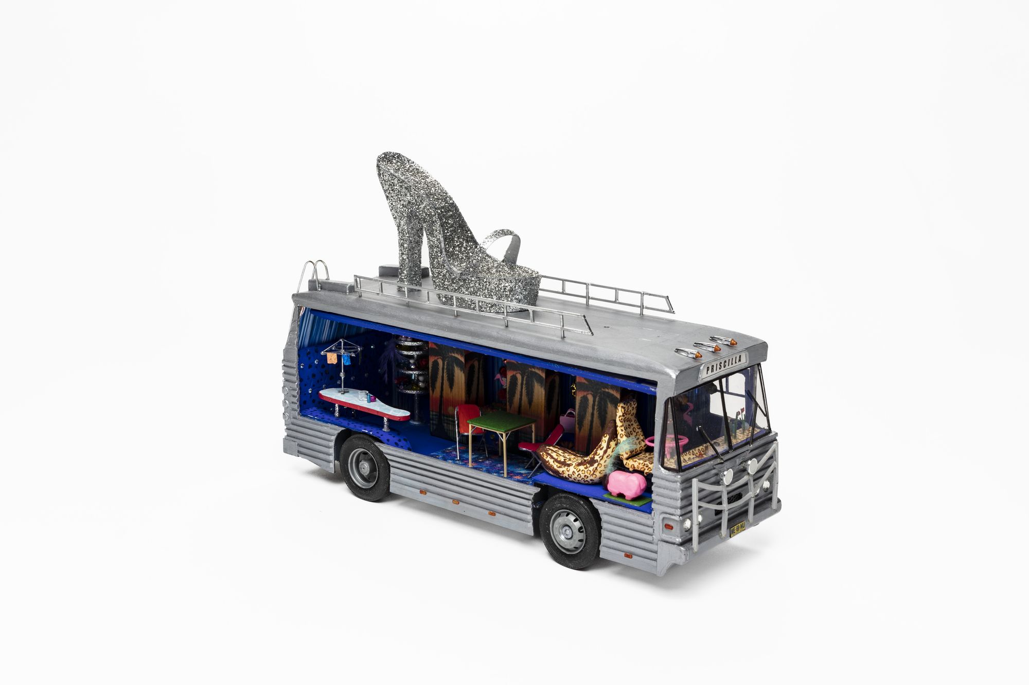 Three quarter side view of silver-coloured model bus with open panel to interior. Inside features brightly coloured miniature furnishings including of a rounded edge bench, hills hoist clothesline, table and two chairs, leopard print covered driver’s seat and banana-shaped lounge, pink sheep and steering wheel. Three panels with an image of a sunset and palm tree hanging on long interior wall of bus. A sign above the windscreen reads ‘Priscilla’ in black lettering. A large silver glittery high heel shoe sits on top of the roof of the bus.