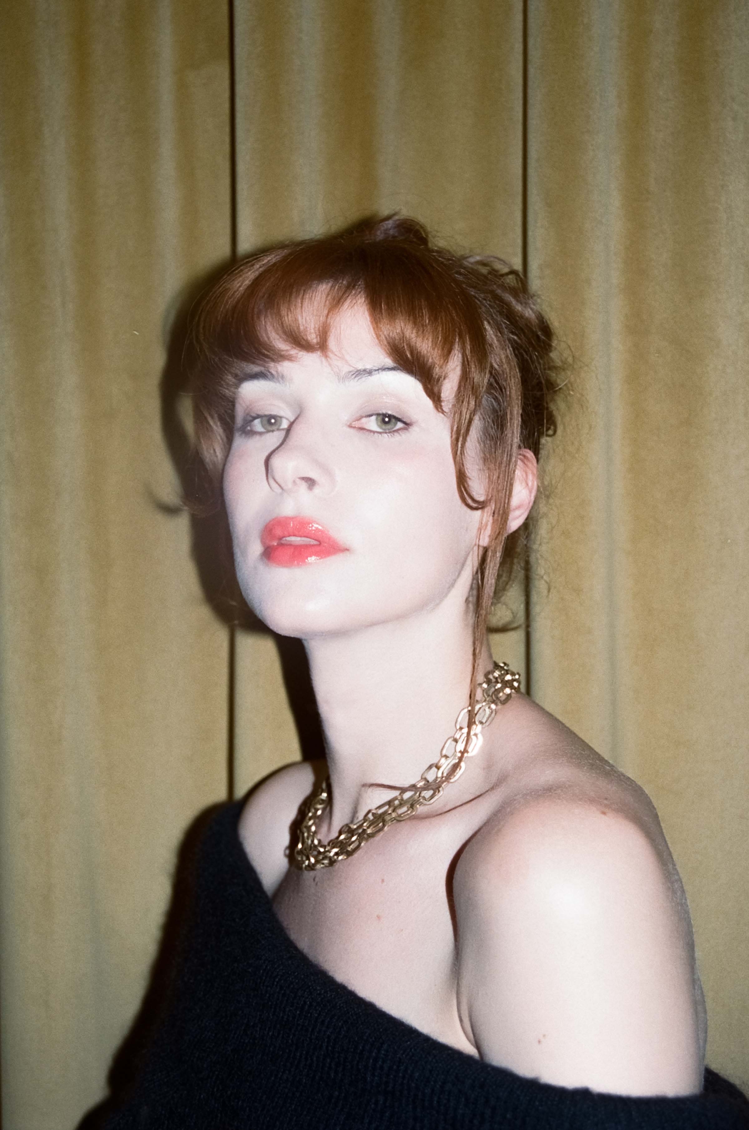 Janet stands in front of a yellow velet curtain, she is wearing a black off the shourlder dress and has thick golden chains around her neck. She looks at us through a beautifully made up face, her red lips just open as if about to smile. Her reddish brown hair is pulled back but there are strands around her face and her curled fringe rests on her eyebrows. The lighting is bright as if this was a moment caught only because of a camera flash.