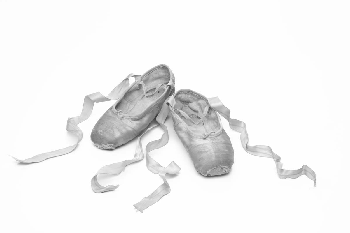 Black and white image of ballet shoes with elastic strap and ribbons curled to side.