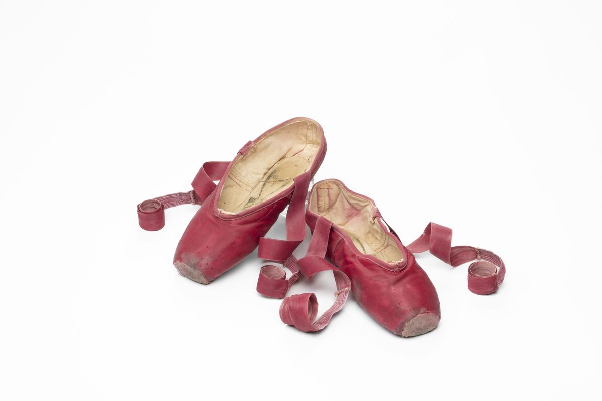 Colour image of worn, red ballet shoes with ribbons curled to the side.