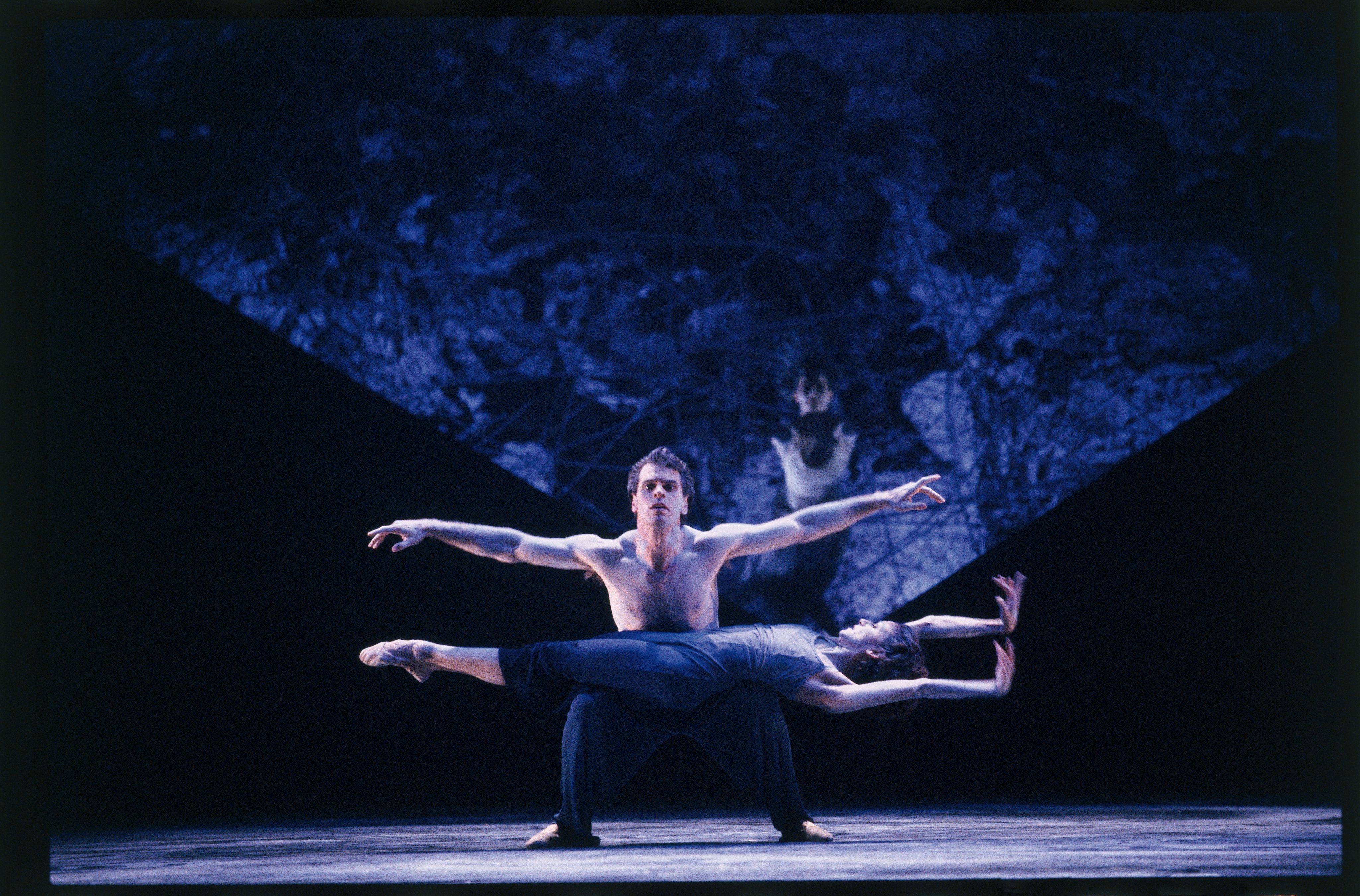 A bare chested man, squatting, with arms outstretched balances a female dancer lying flat on his knees. In the background, a female dancer looms.
