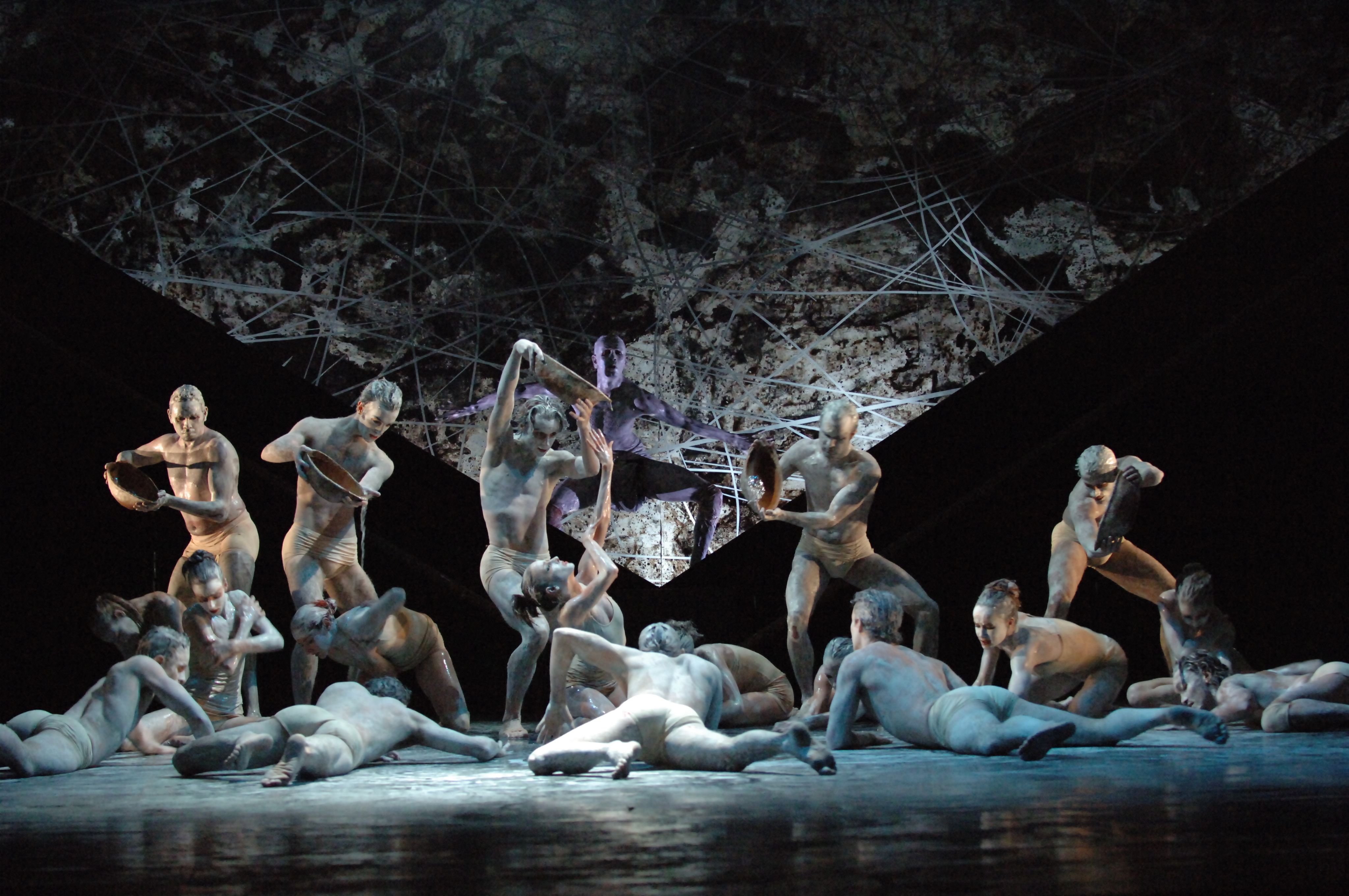 A group of white-painted dancers in varying degrees of lying and standing on a stage. Five hold vessels dripping liquid on to dancers below.