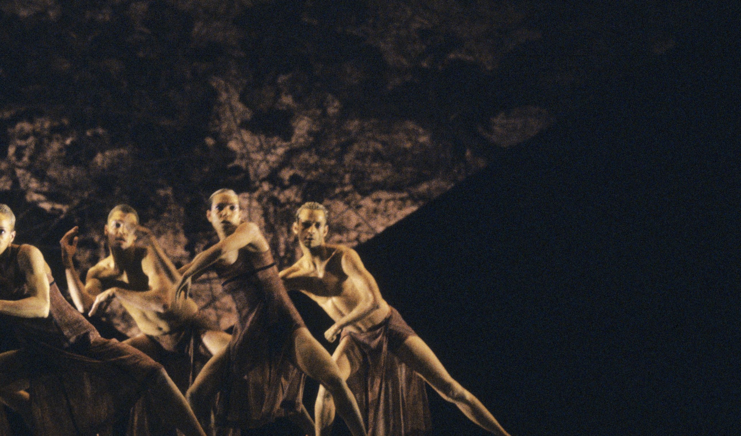 A group of male and female dancers on stage, leaning backward with arms at right angles.