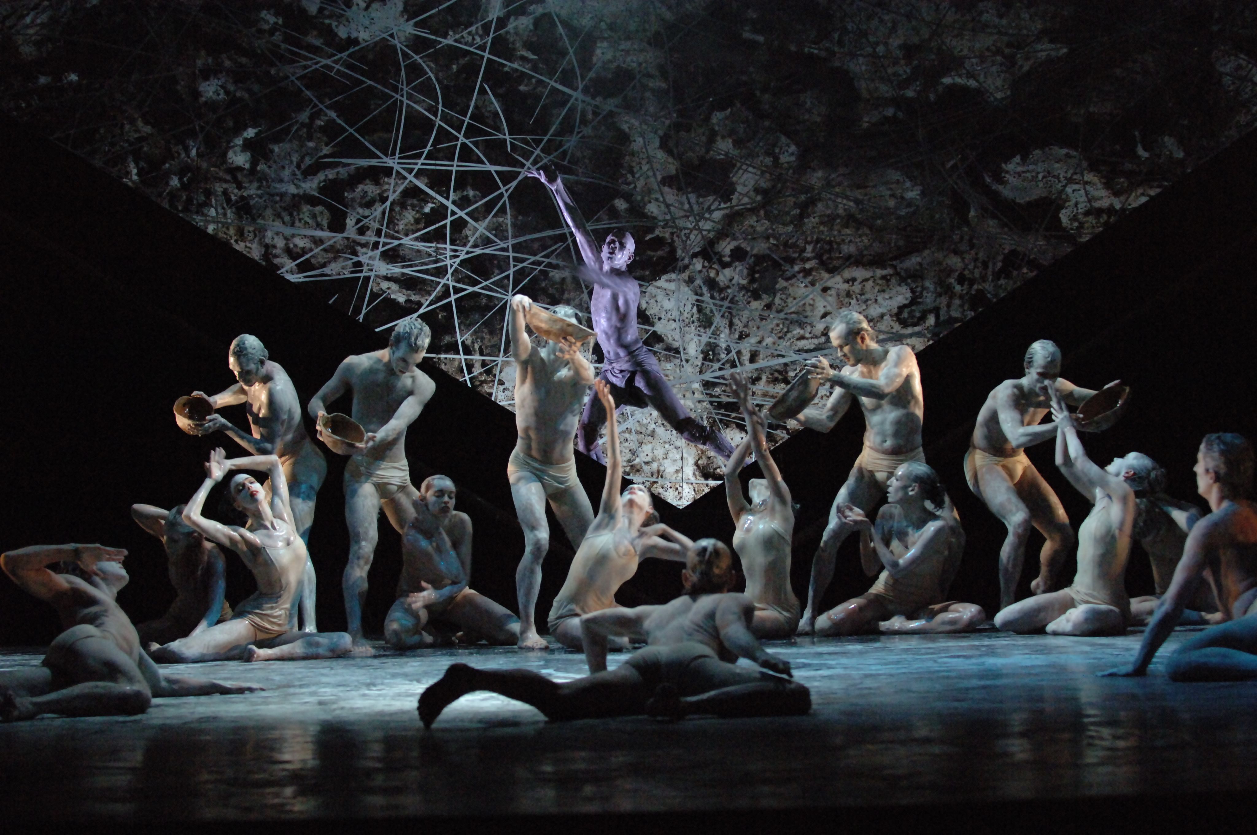 A group of white-painted dancers in varying degrees of sitting and standing on a stage. Five hold vessels dripping liquid on to dancers below.