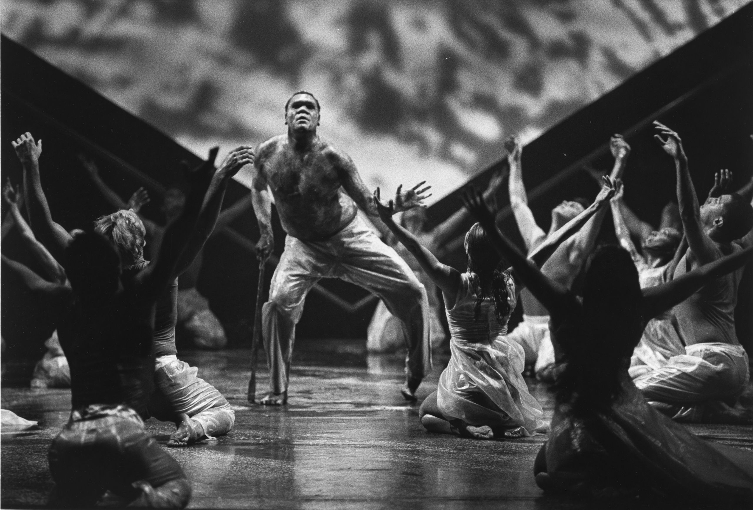 Bare-chested male dancer on stage leans on a walking stick. Around him other dancers are seated with arms raised in a V shape.