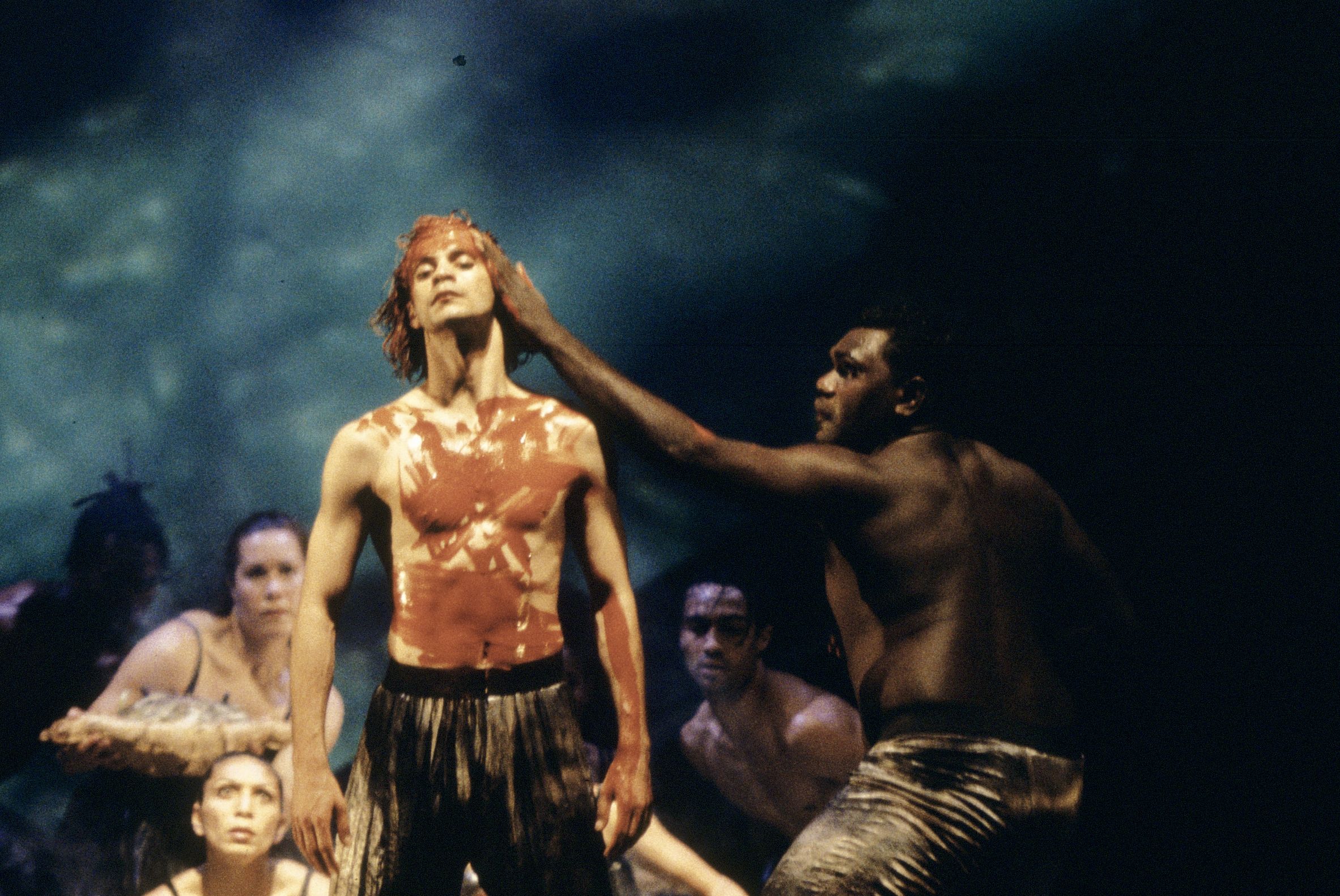 Two bare chested males on stage. One is leaning in with his hand on the others face, which features red, painted markings along with his chest and arms. A group of dancers crouch behind them.