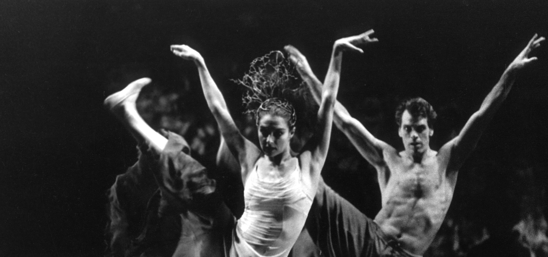 A female dancer wearing a crown stands on stage with arms and one leg in the air. A male dancer in the background holds the same position.