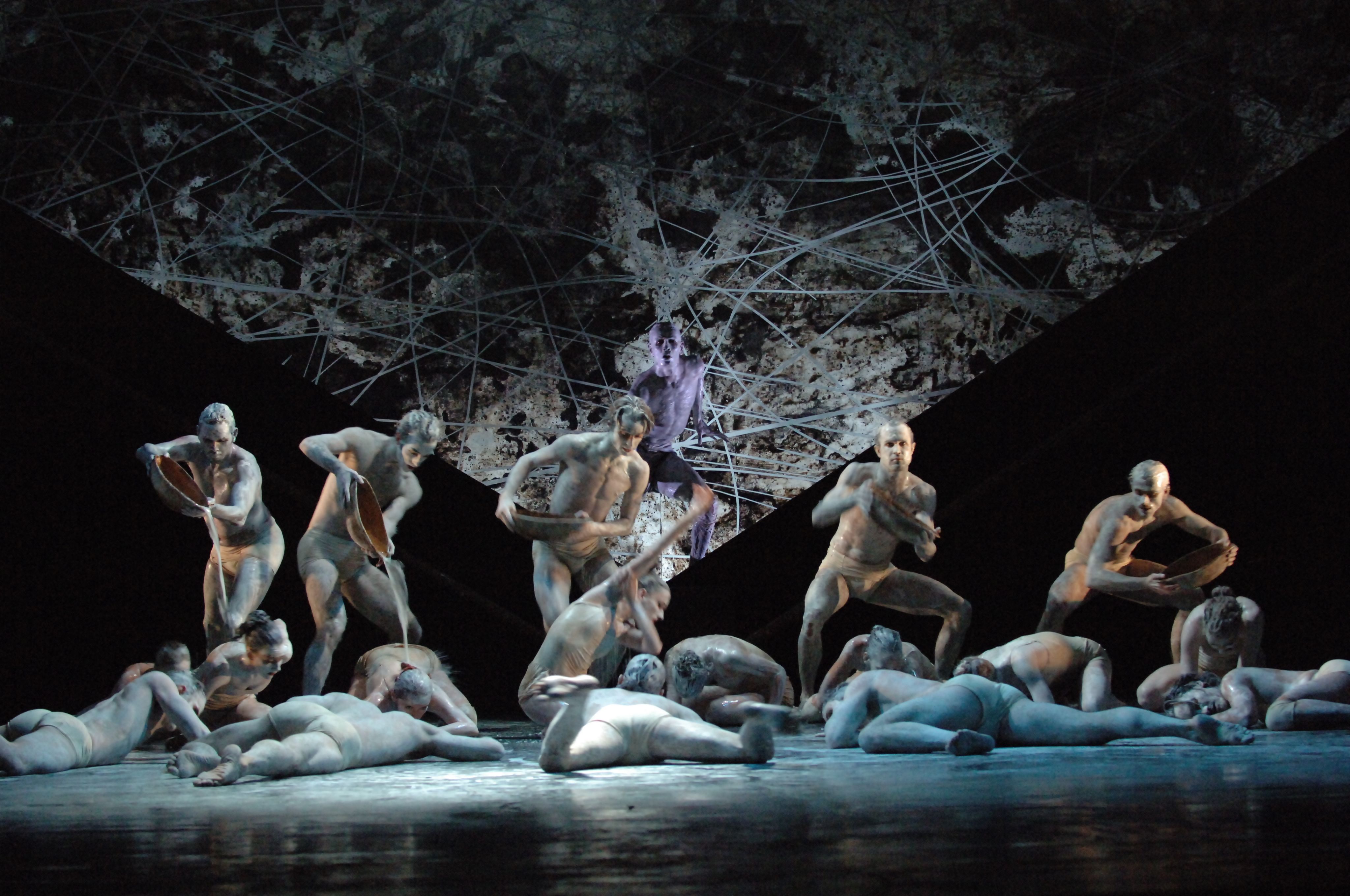 A group of white-painted dancers in varying degrees of lying and standing on a stage. Five hold vessels dripping liquid on to dancers below.