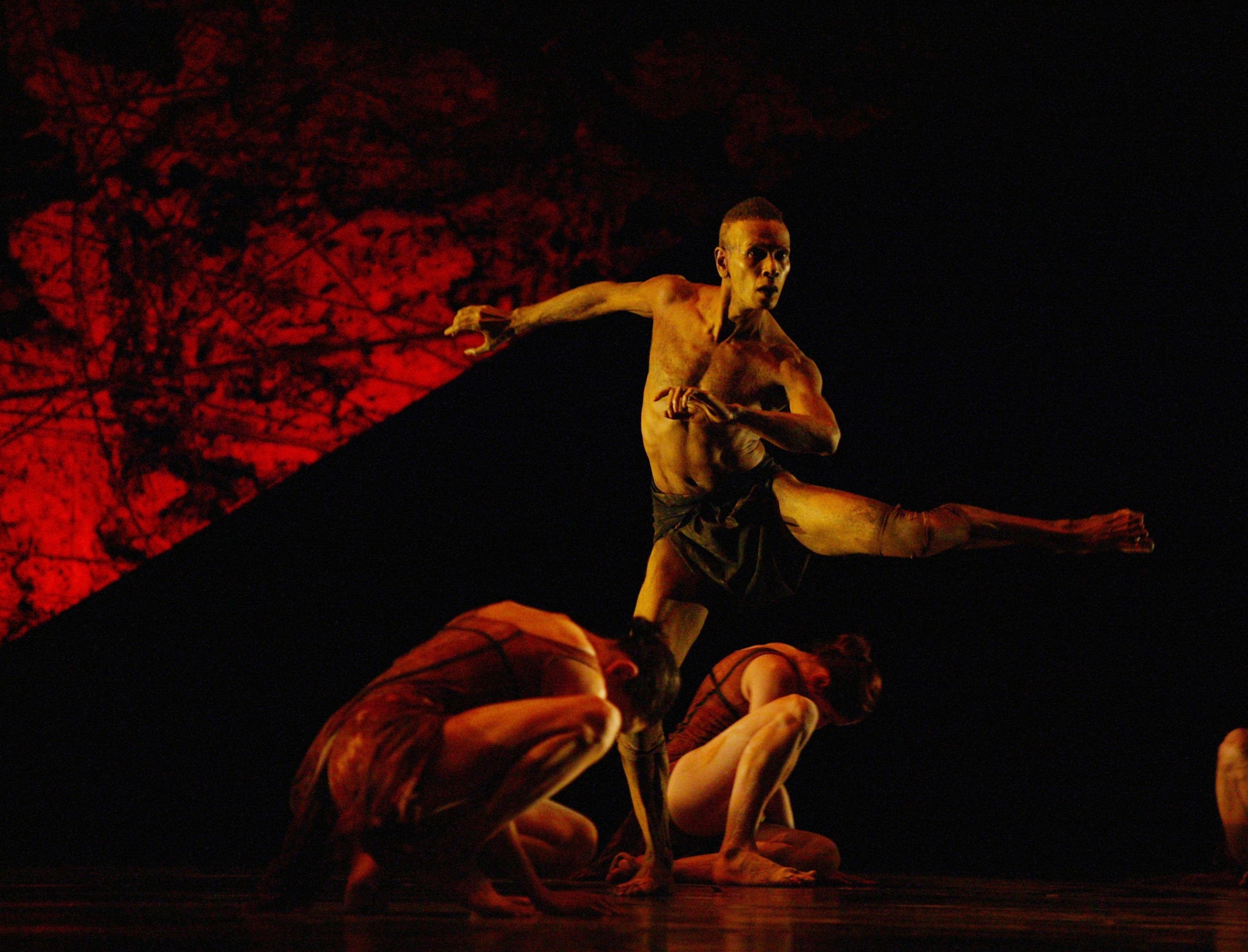 wo female dancers crouch on stage floor. A male dancer is between them with one leg outstretched.