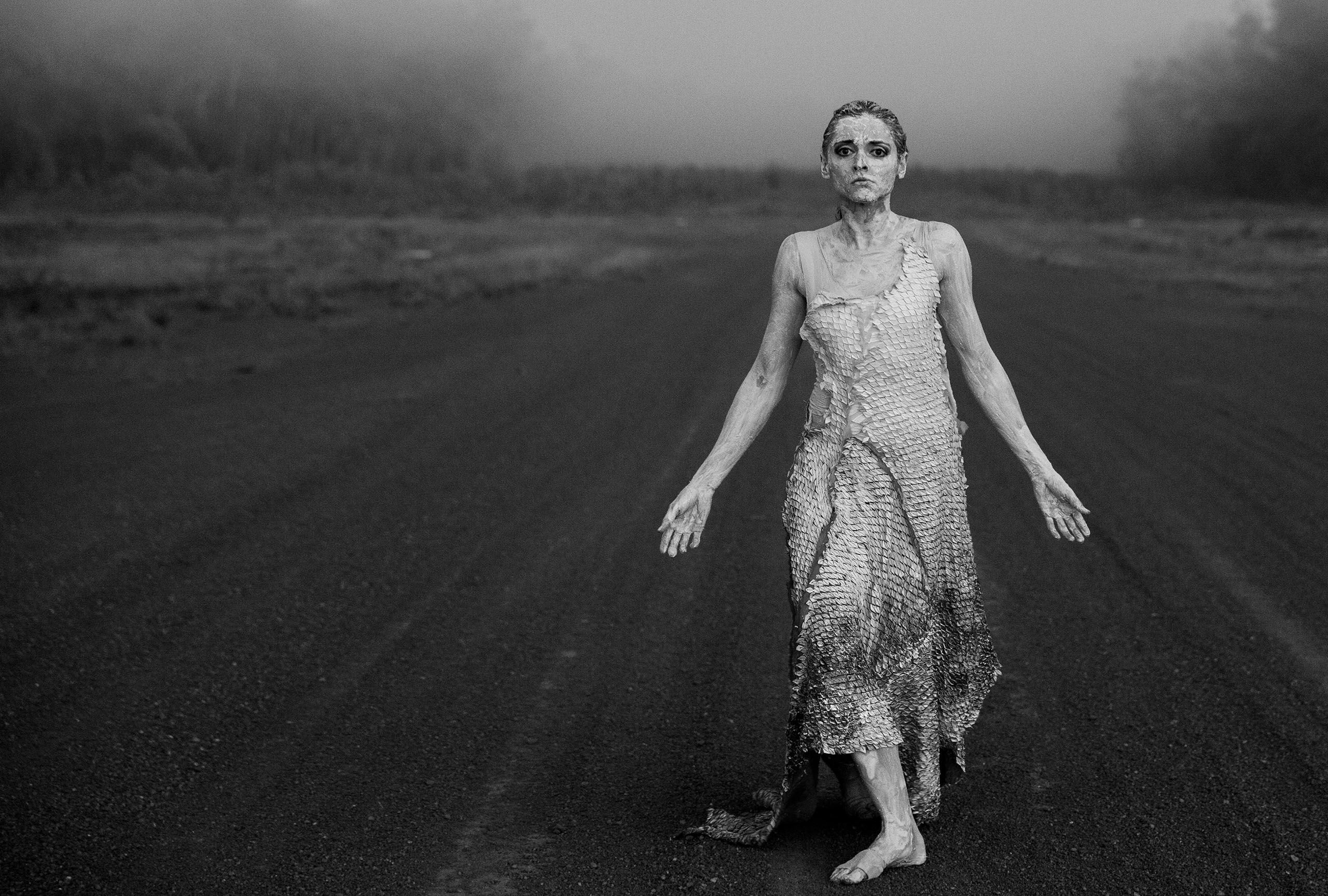 Woman with arms outstretched walks along deserted country road. She has white ochre on her face and body.