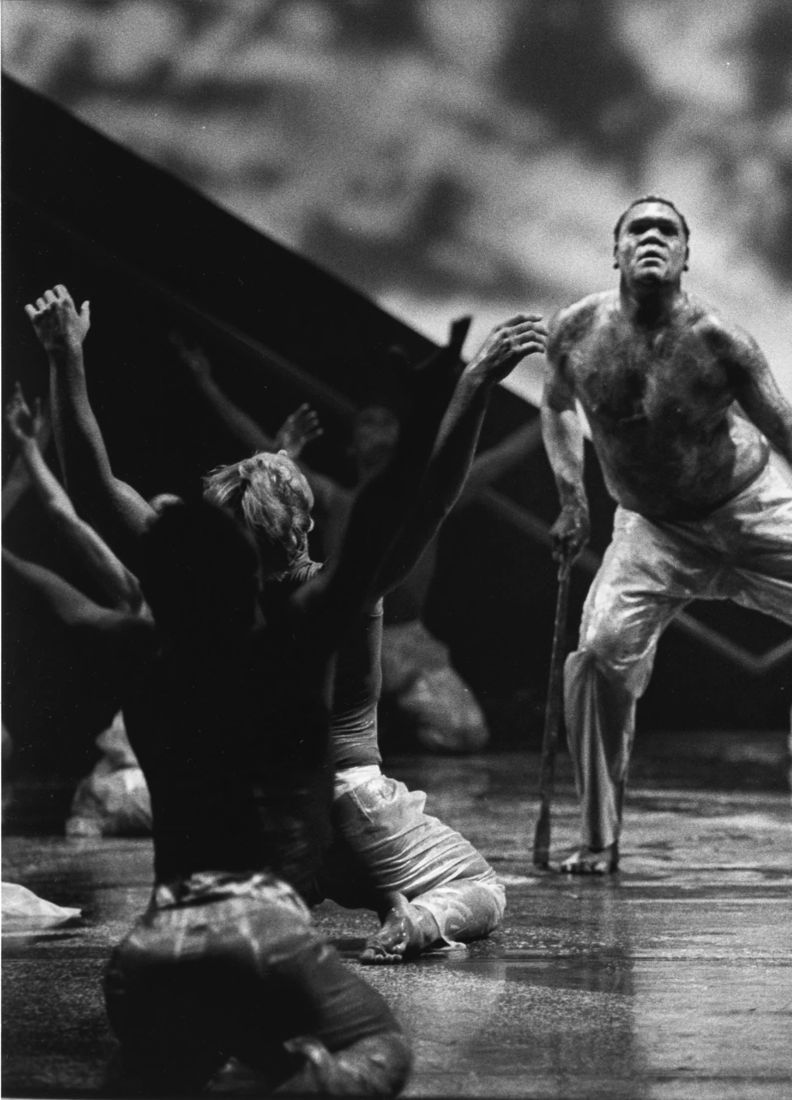 Bare-chested male dancer on stage leans on a walking stick. Around him other dancers are seated with arms raised in a V shape.