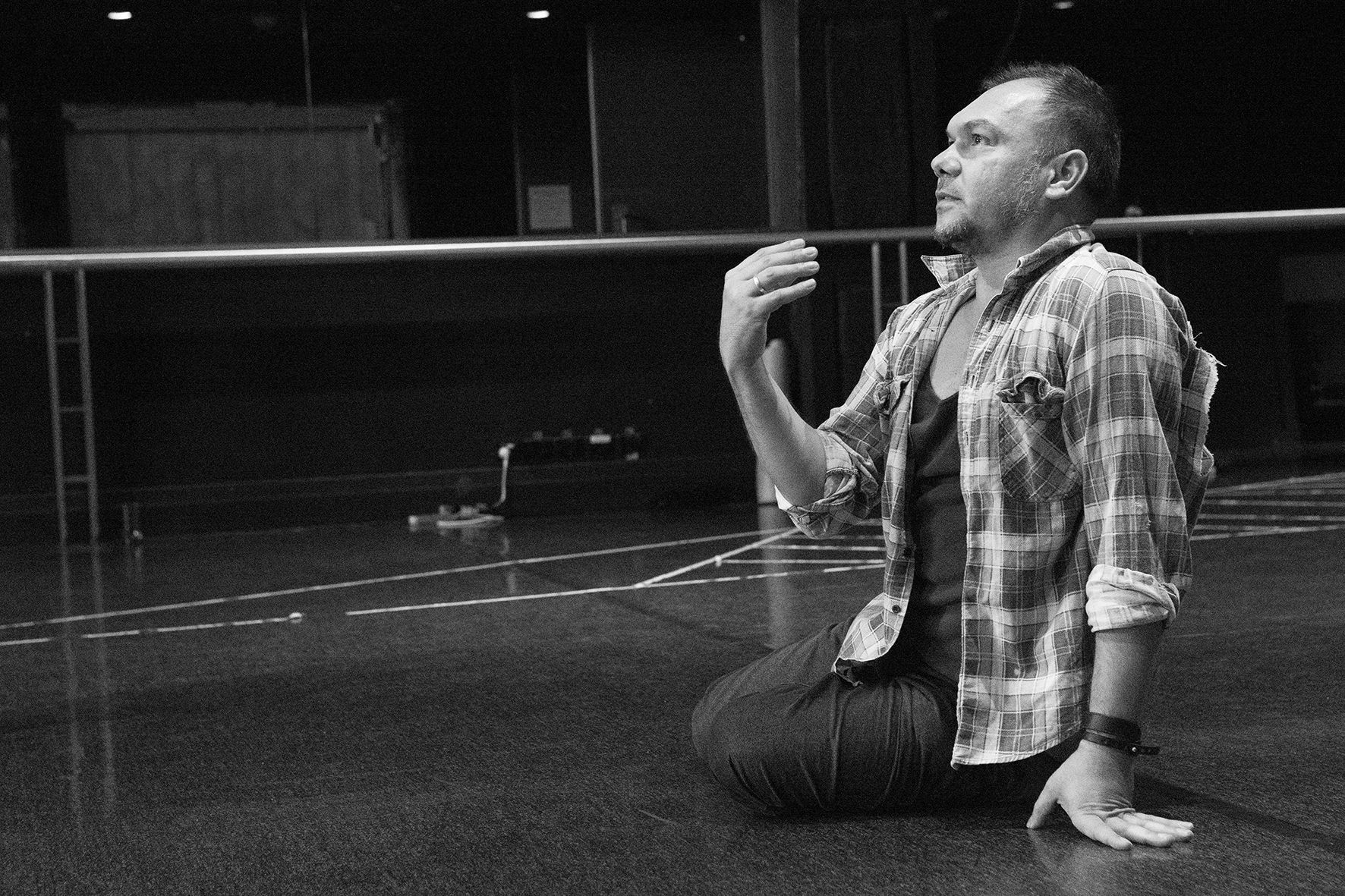 Man sitting on floor of dance studio, gestures with his right hand as though explaining something.