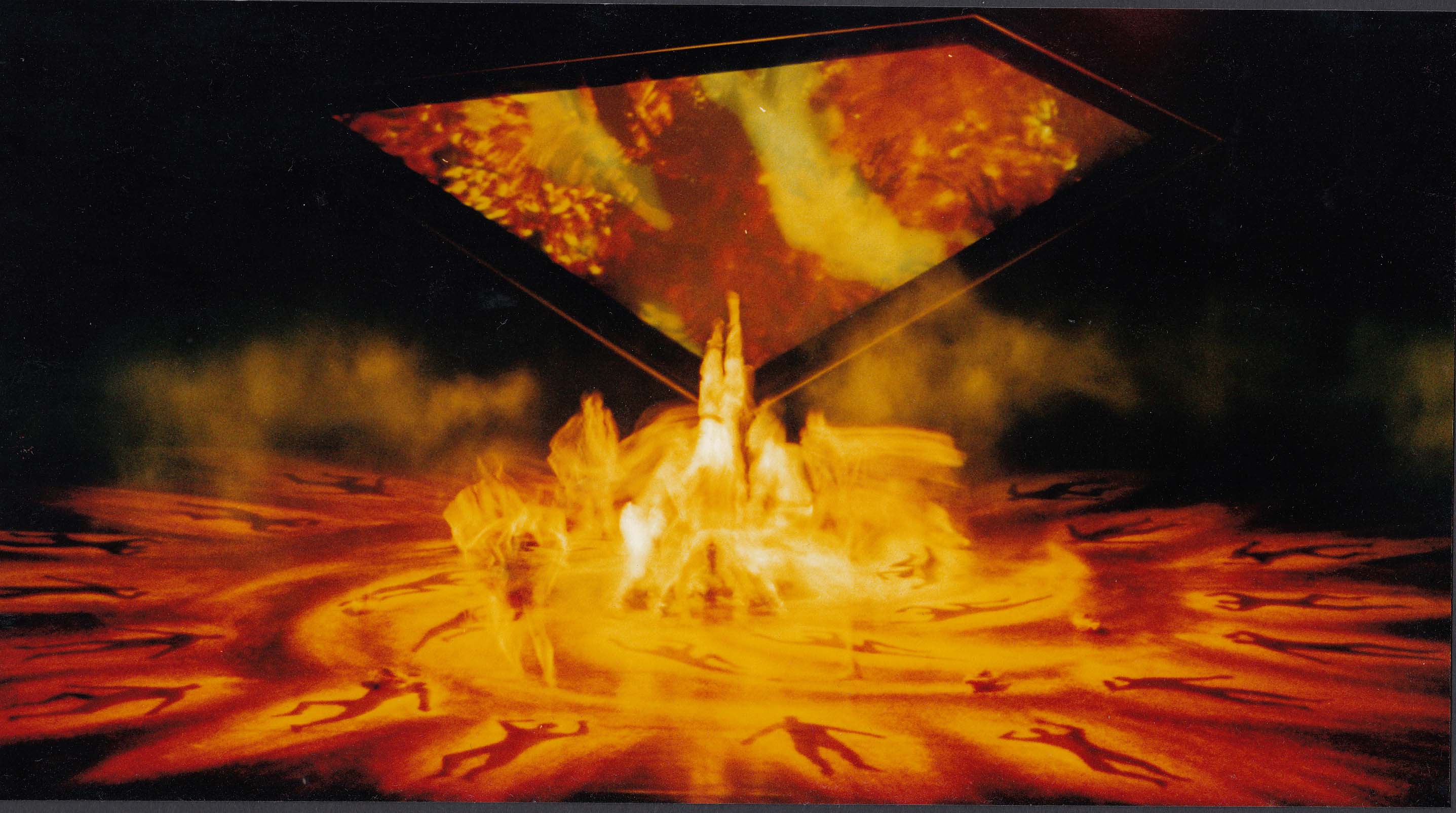 Time lapse, wide shot of stage featuring a group of dancers' movements highlighted in orand lighting. Orange outlines of bodies decorate the floor in a circle around the dancers.