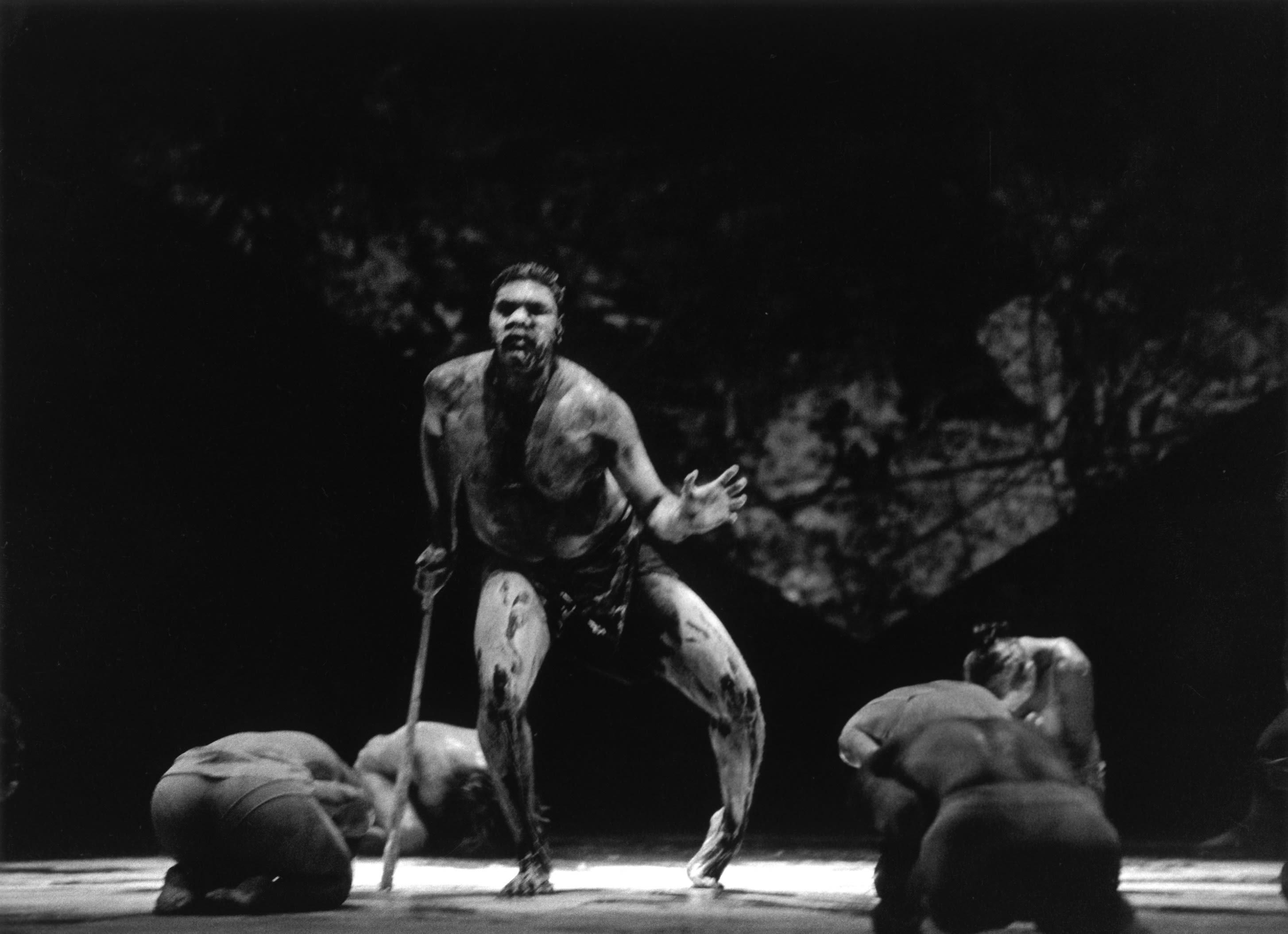Bare-chested male dancer on stage leans on a walking stick. A group of dancers crouch around him, looking down.
