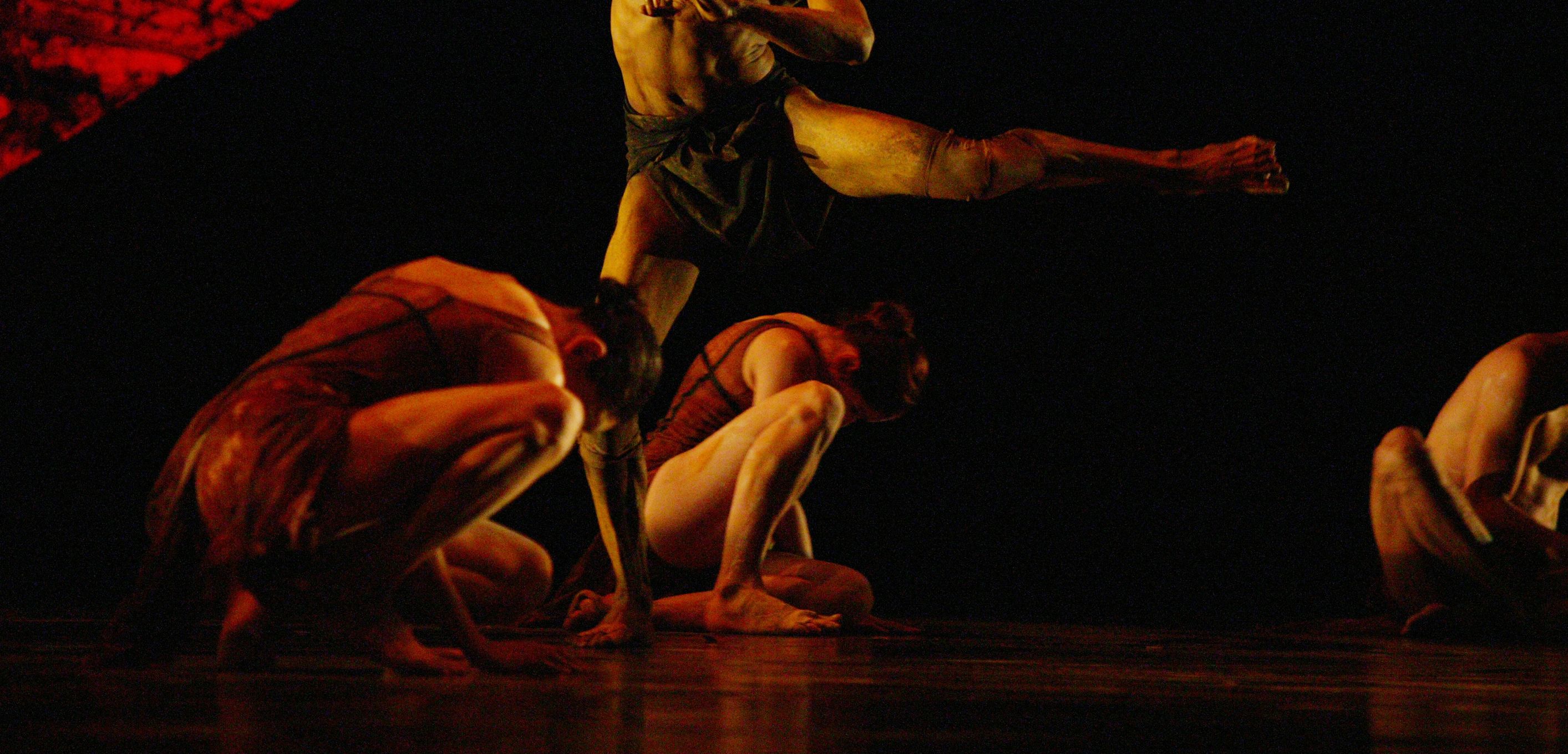 Two female dancers crouch on stage floor. A male dancer is between them with one leg outstretched.
