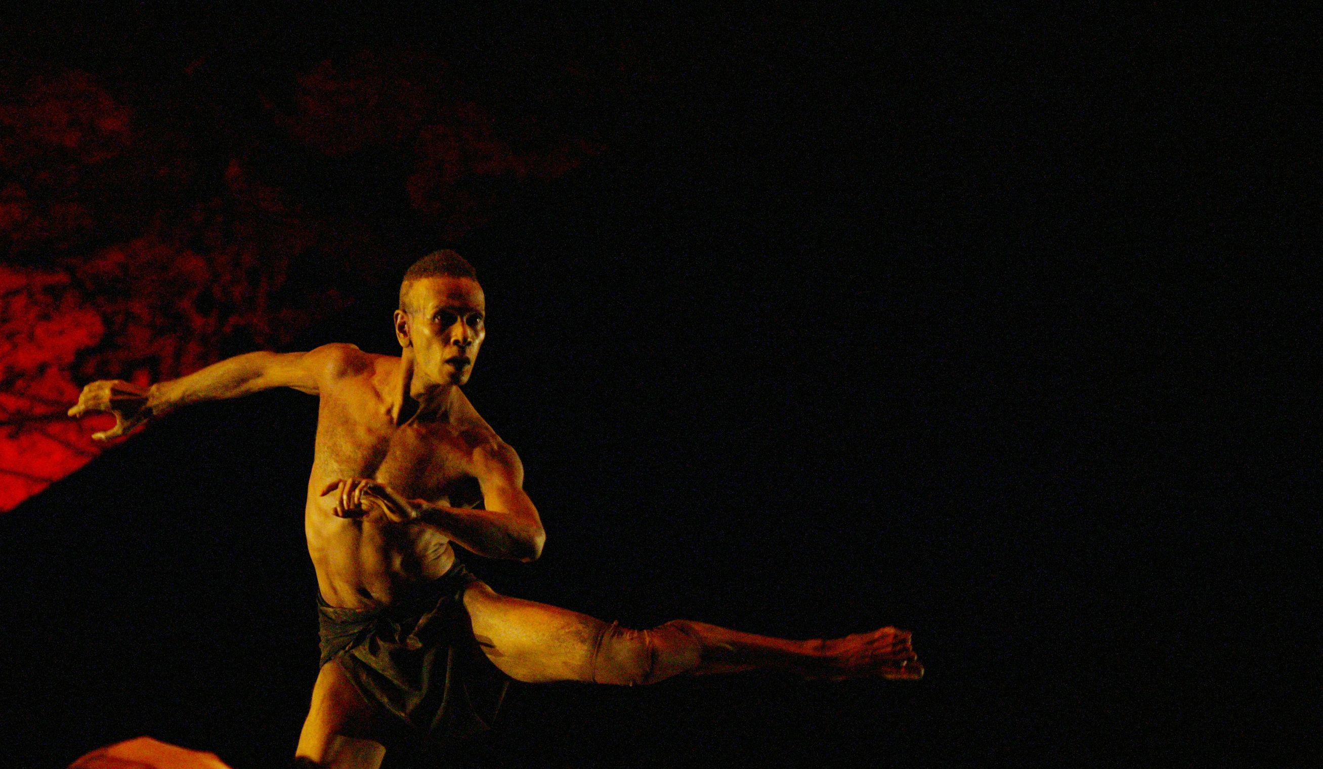 Bare-chested male dancer on stage looks forward with one leg outstretched.