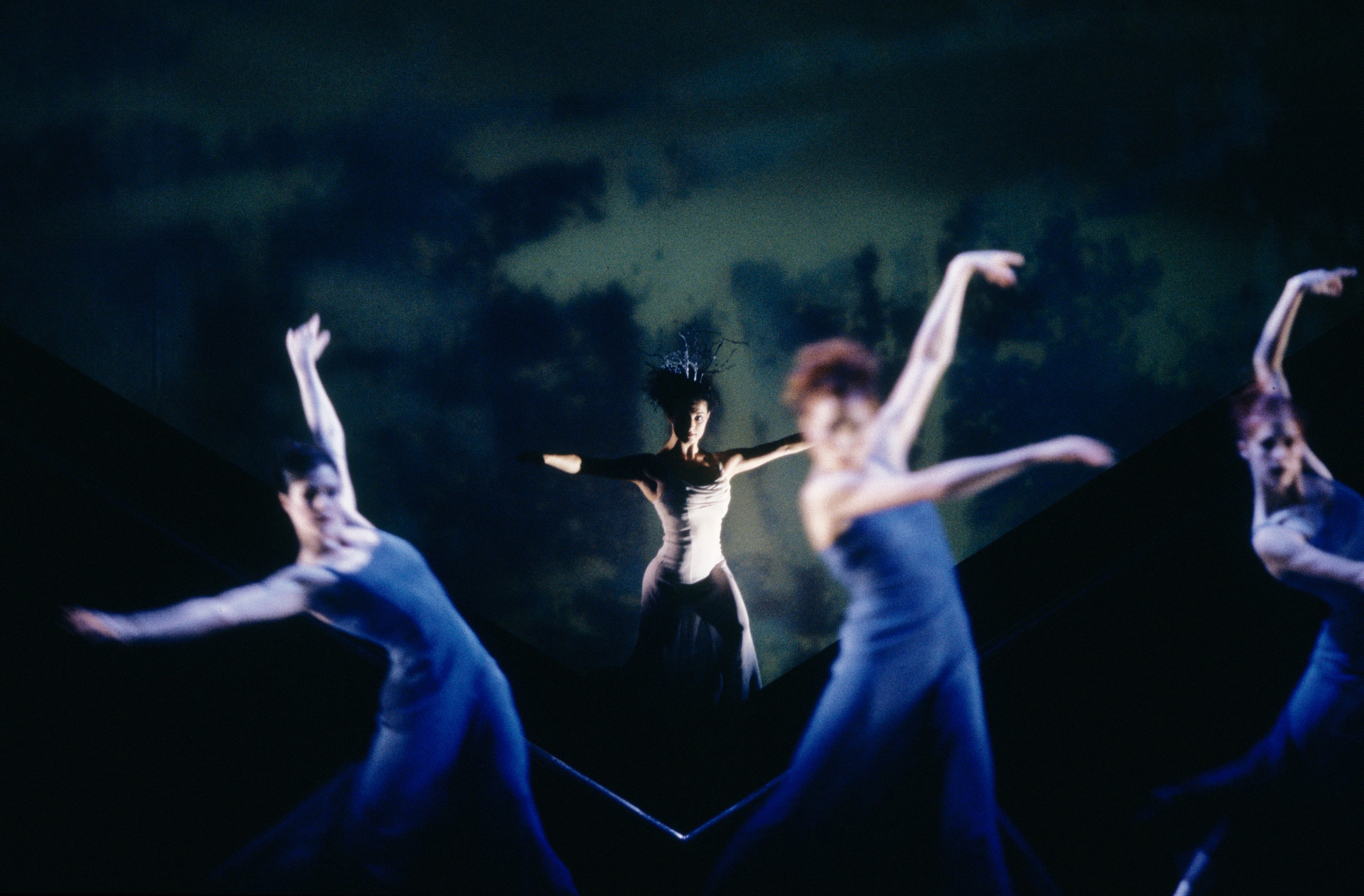 A female dancer, wearing a crown, stands with arms outstretched. Three dancers are in montion in the foreground.