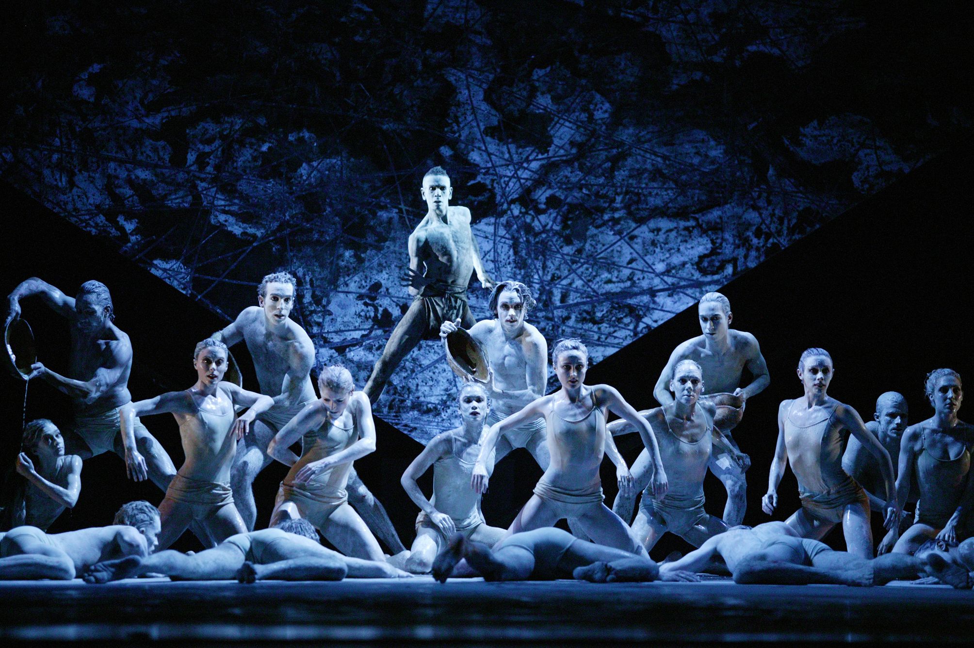 A group of white-painted dancers in varying degrees of sitting and standing on a stage. Five hold vessels dripping liquid on to dancers below.