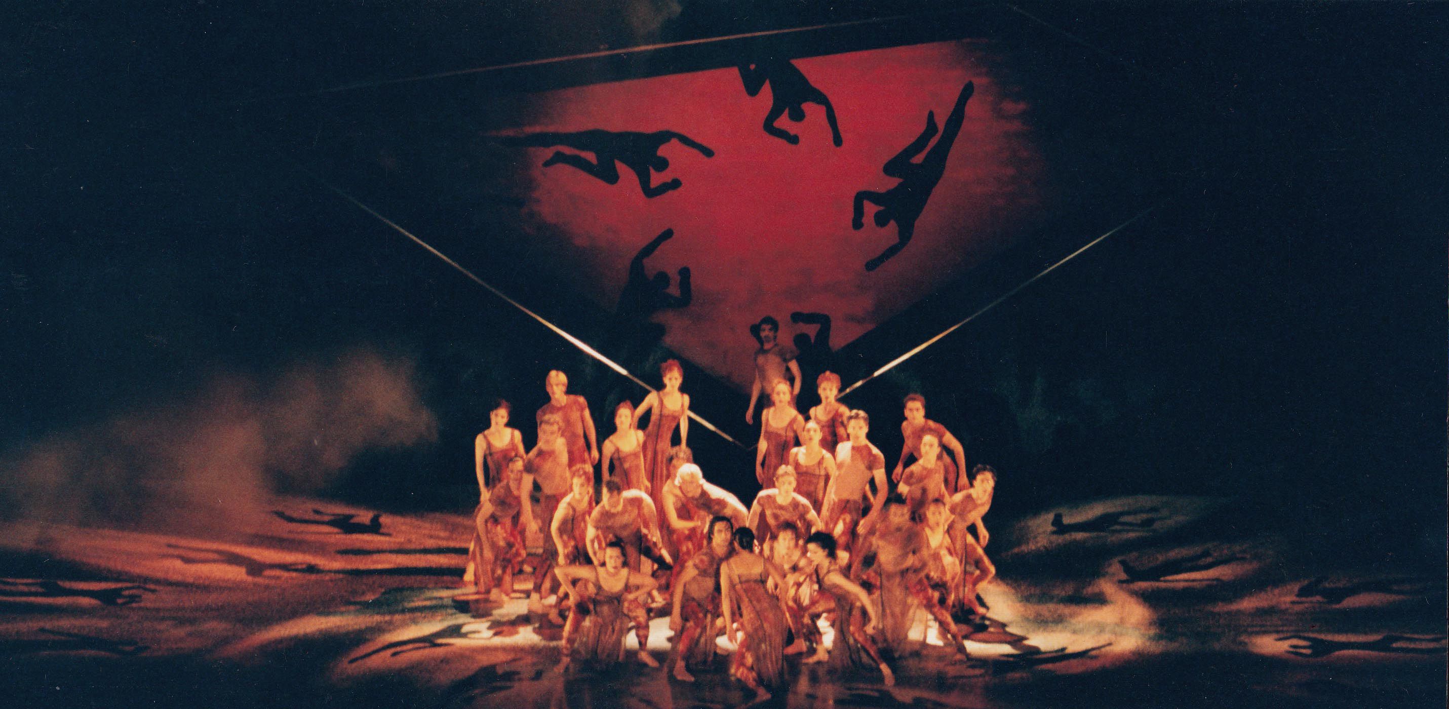 Large group of dancers gathered in the centre of a stage. Lighting and backdrop are red.