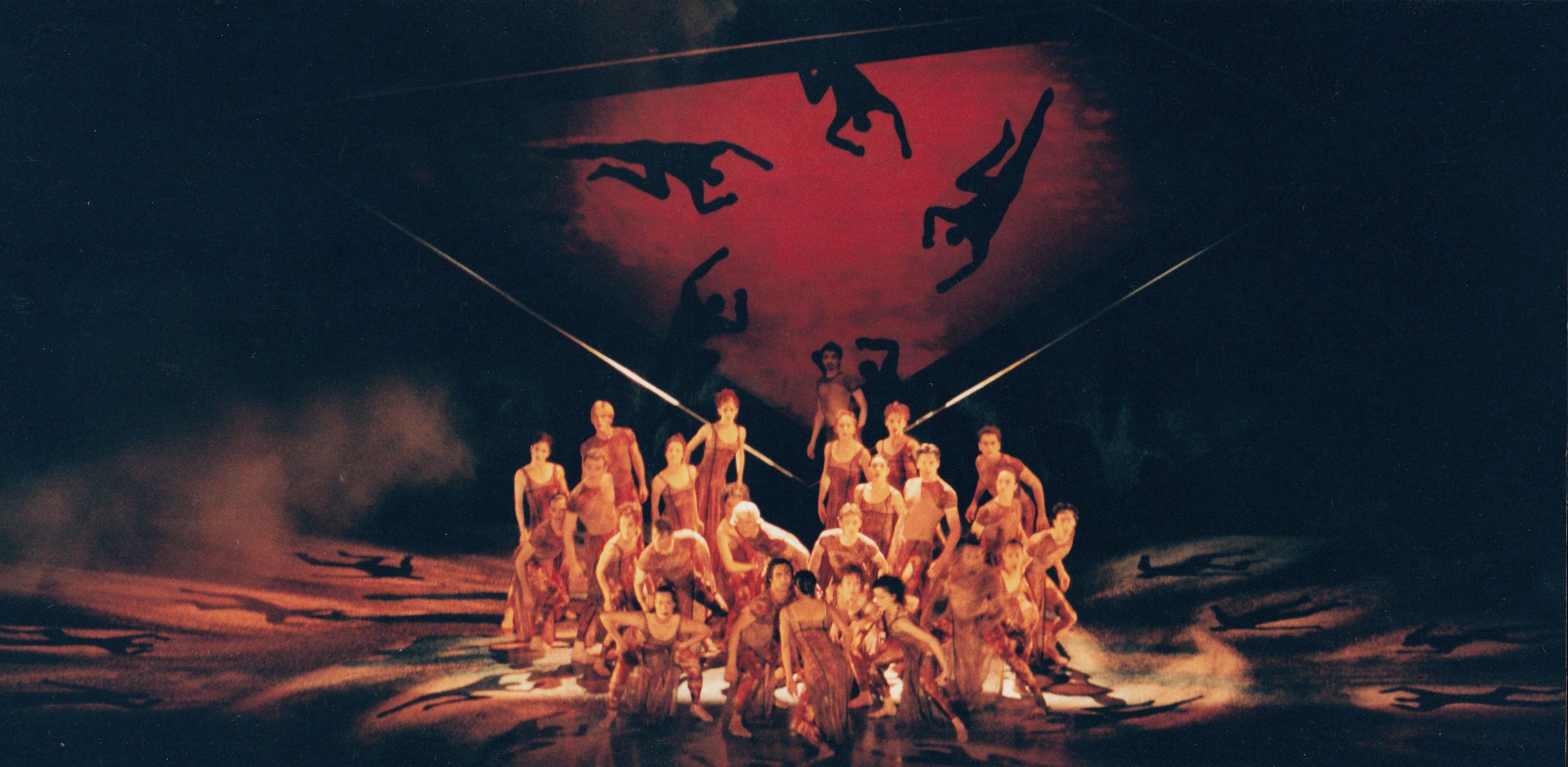 Large group of dancers gathered in the centre of a stage. Lighting and backdrop are red.