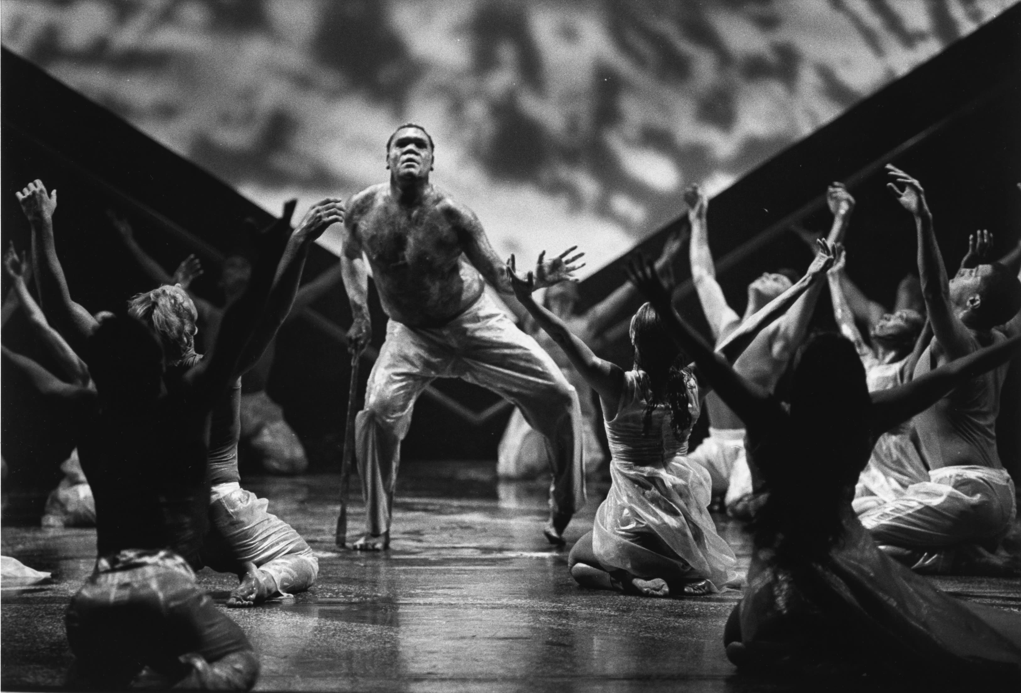 Bare-chested male dancer on stage leans on a walking stick. Around him other dancers are seated with arms raised in a V shape.