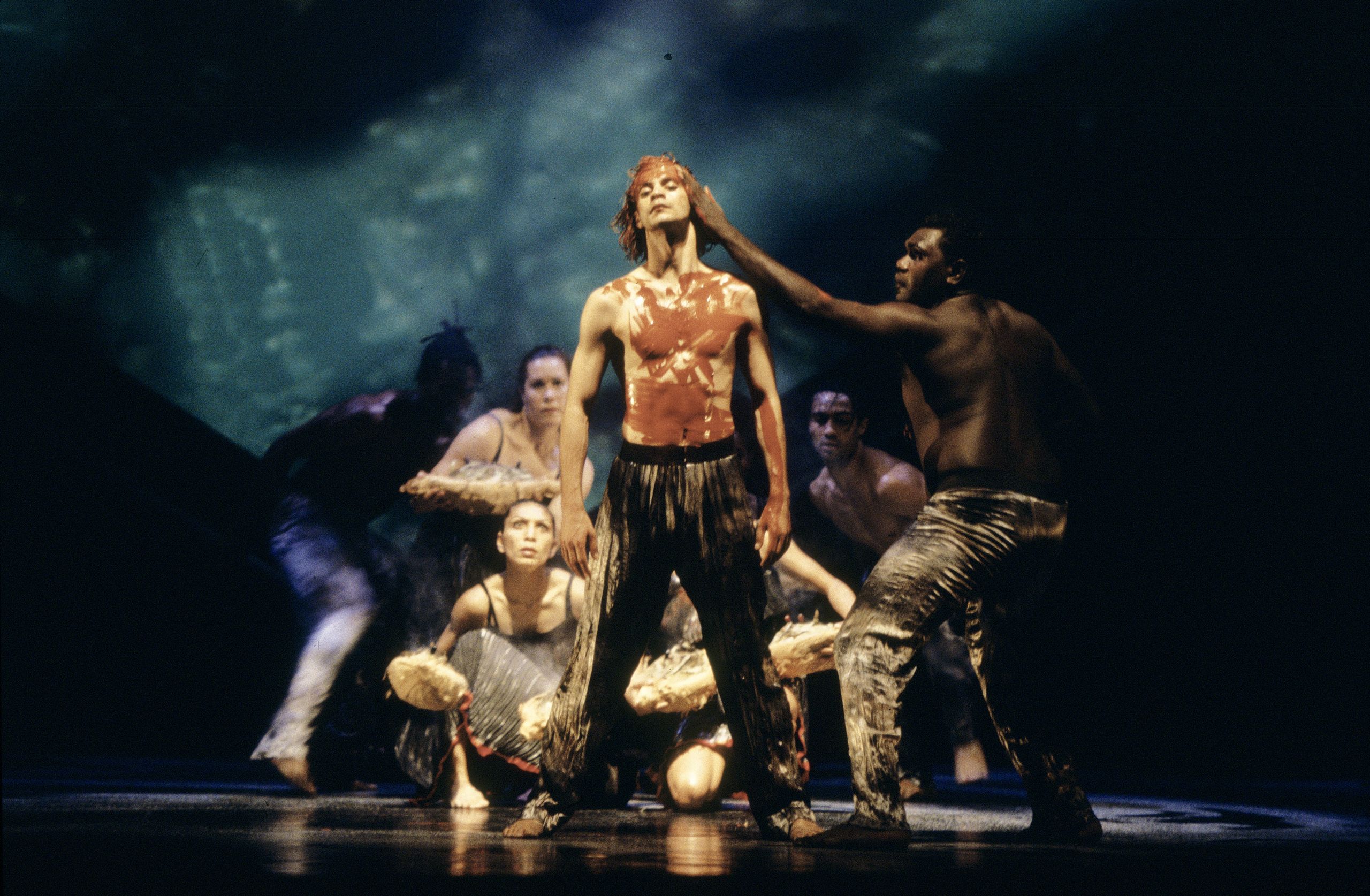 Two bare chested males on stage. One is leaning in with his hand on the others face, which features red, painted markings along with his chest and arms. A group of dancers crouch behind them.