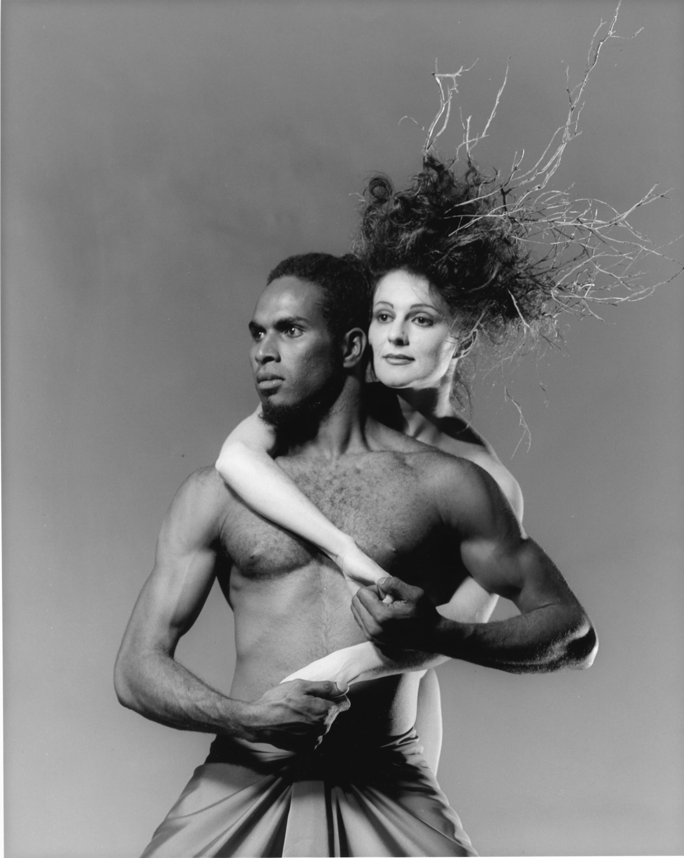 Close up of head and chest of man, looking to the left, with a woman behind who is draping her arms around his shoulders. They hold hands.