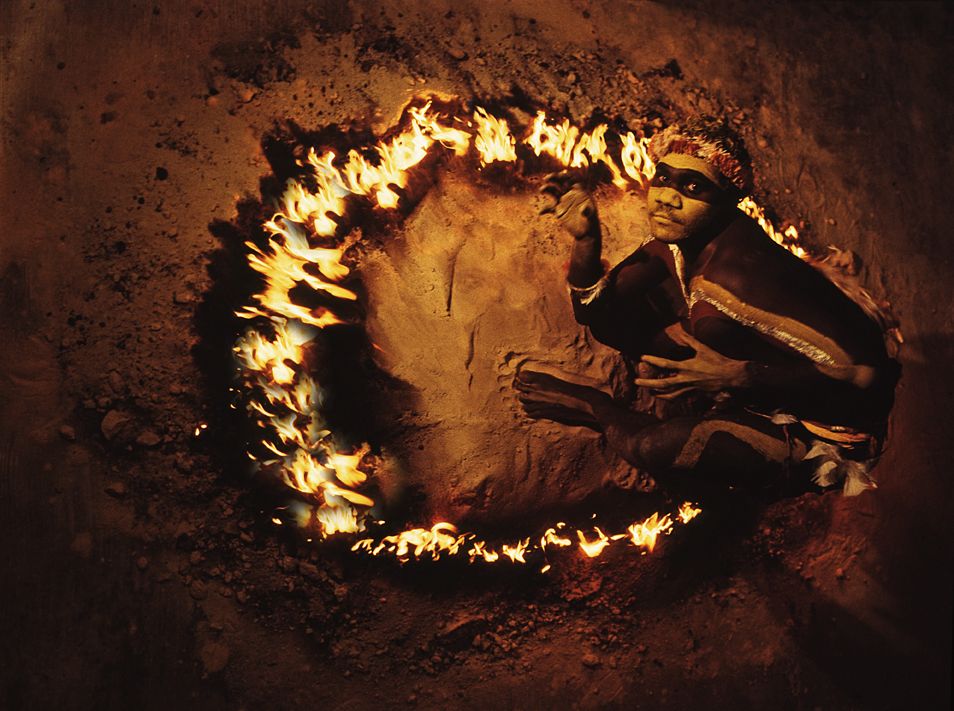 A man with white ochre on his face and body crouched beside a ring of fire situated on a bed of sand. He is looking towards the camera and his hands mimic claws.