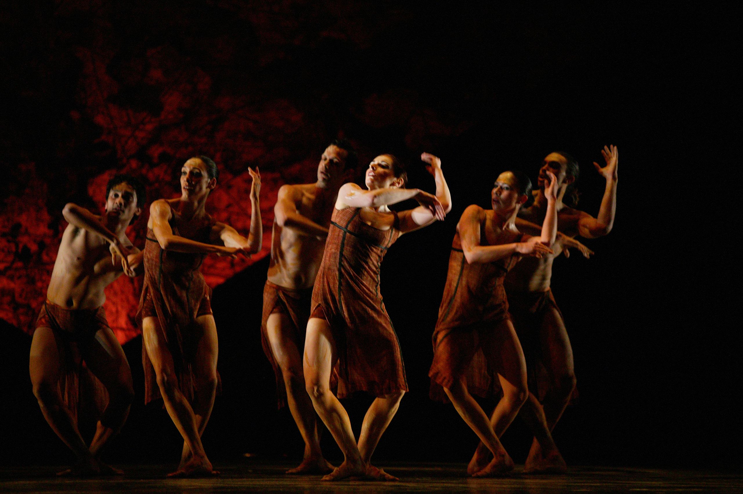 Large group of dancers gathered in the centre of a stage, with a black and red backdrop..