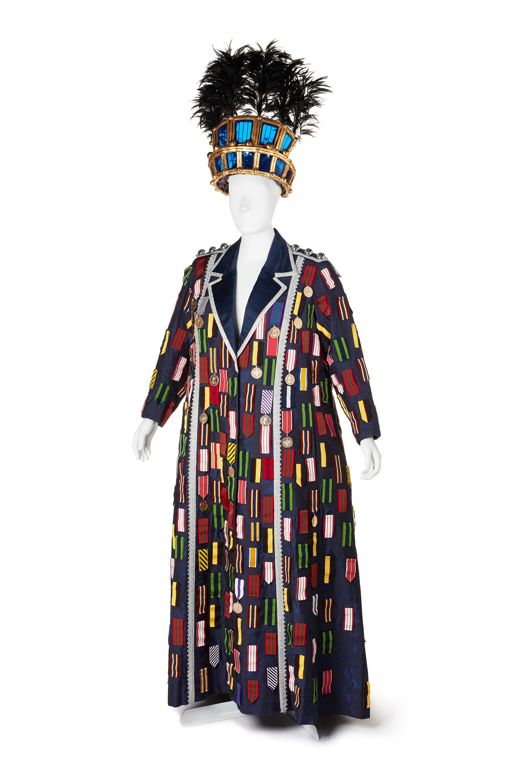 Photograph of a woman's costume on a white mannequin. It features a navy blue, floor lenght coat with medals all over. A headdress of gold and blue is topped with black feathers.