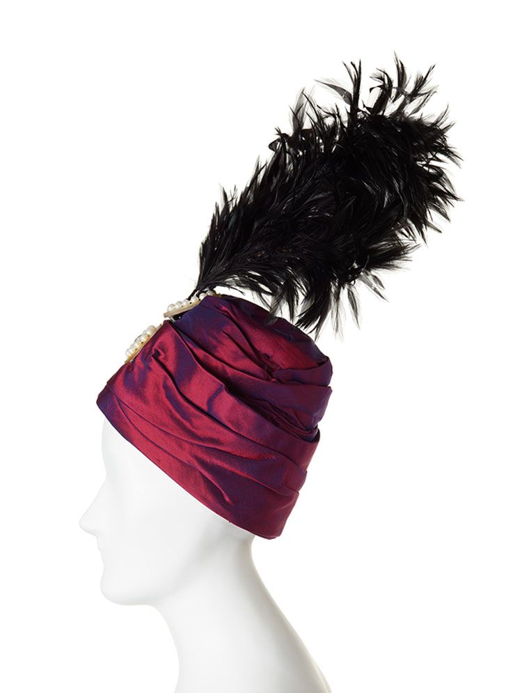 Side, close-up of headdress on white mannequin. It is a turban-like hat in purple mettalic fabrics with jewels at the fron and a plume of black feathers stems from the top.