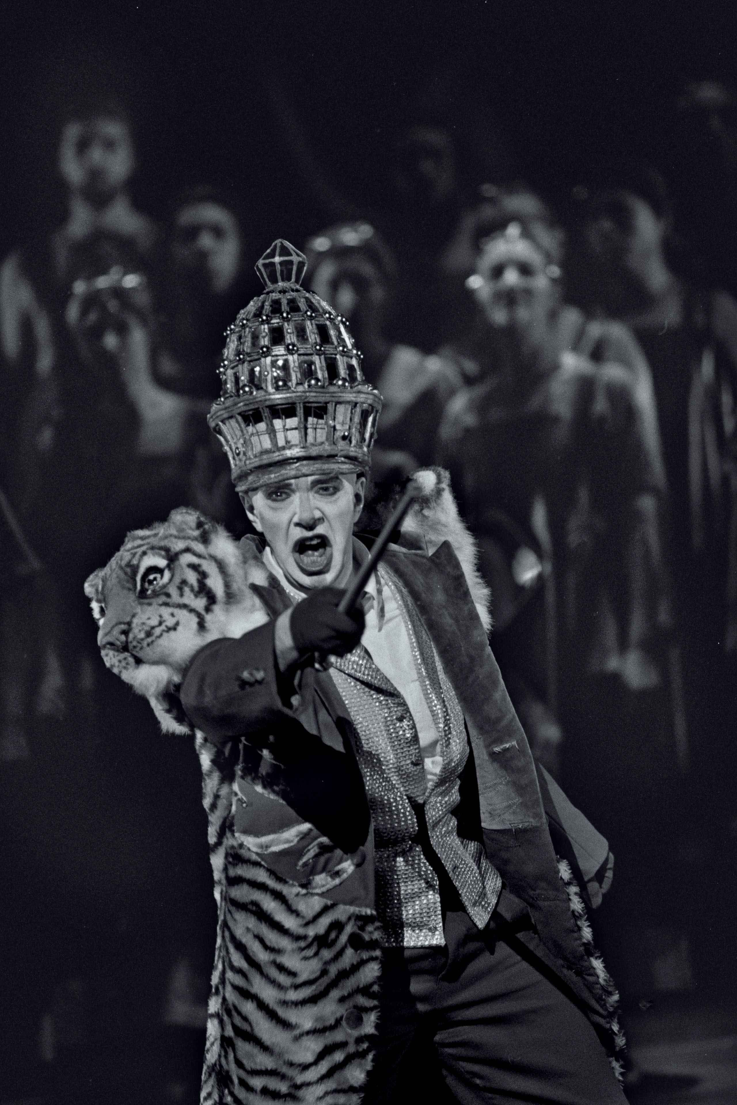 Black and white photograph of performer on stage wering a costume featuring a 3D tigers head on the shoulder and a headdress. He has his face painted white and is pointing a cane outwards.