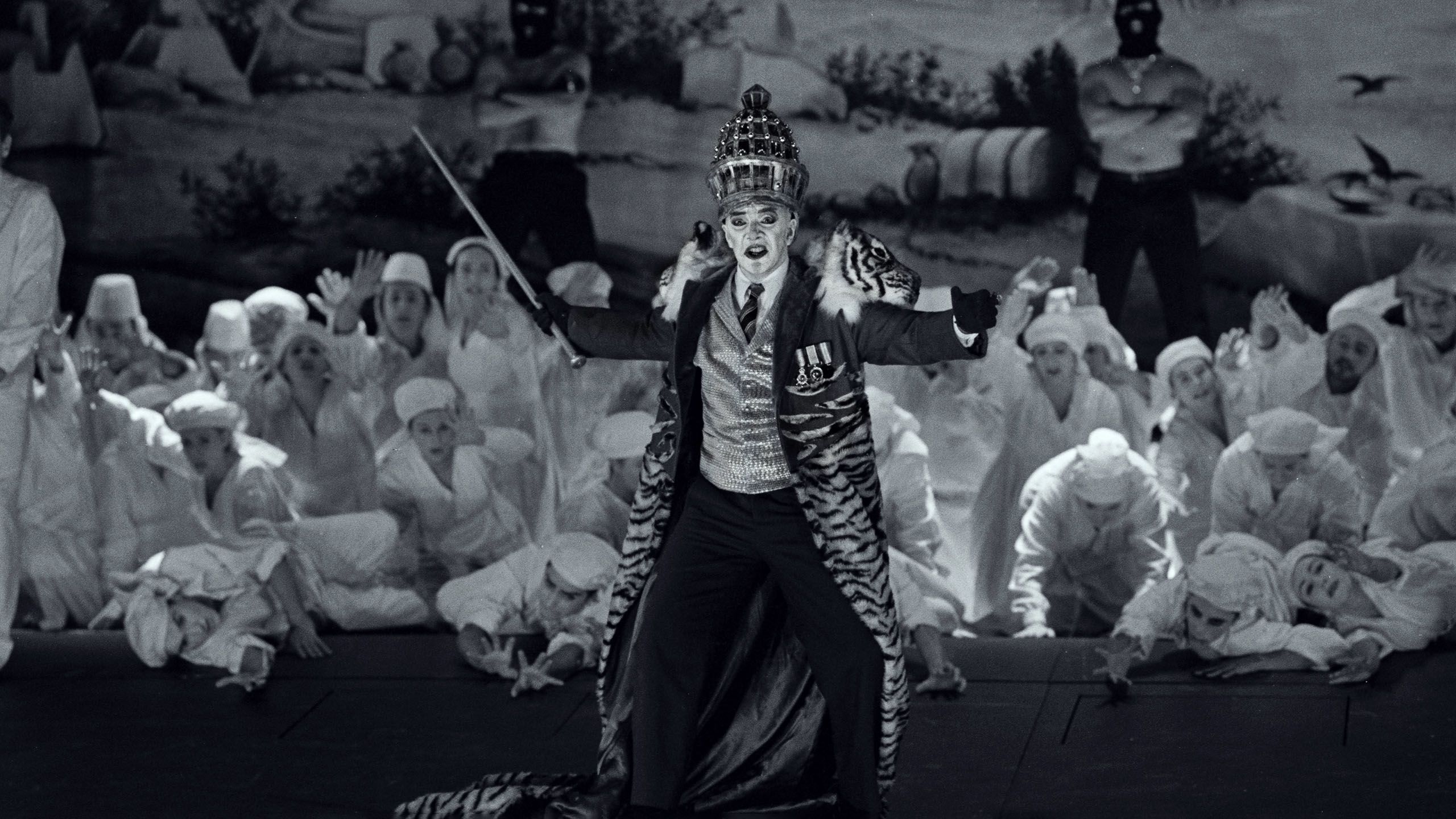 Black and white photograph of theatre stage. A group of actors in white fill the background and a single actor in an eloborate costume and headdress gestures with arms outstretched.