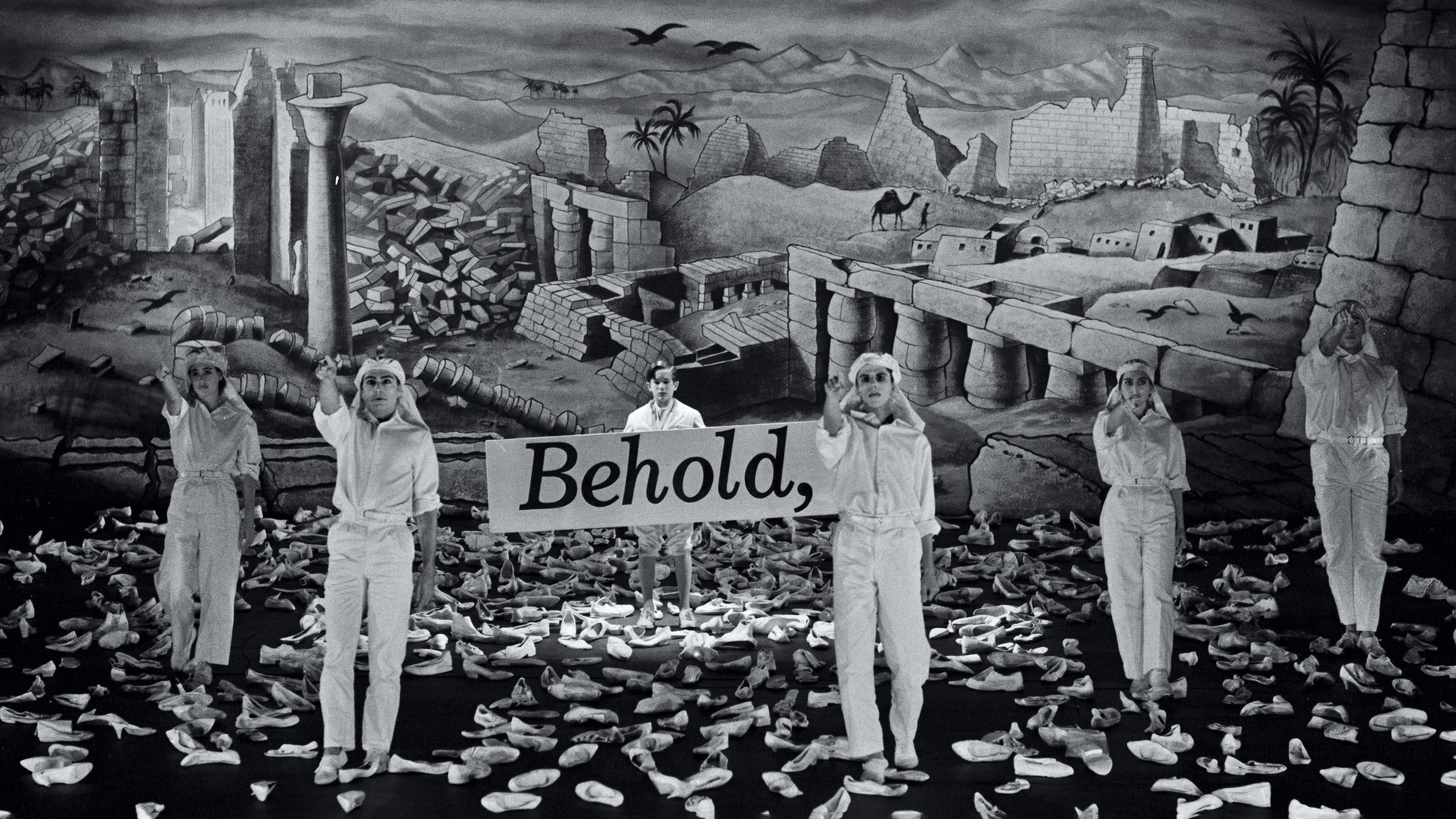 Black and white photograph of theatre stage. The background features a landscape of ruined stone buildings. On stage six actors in white stand and point towards the audience, one holds a sign written with 'Behold,'. They are surrounded by a sea of white shoes on the floor.
