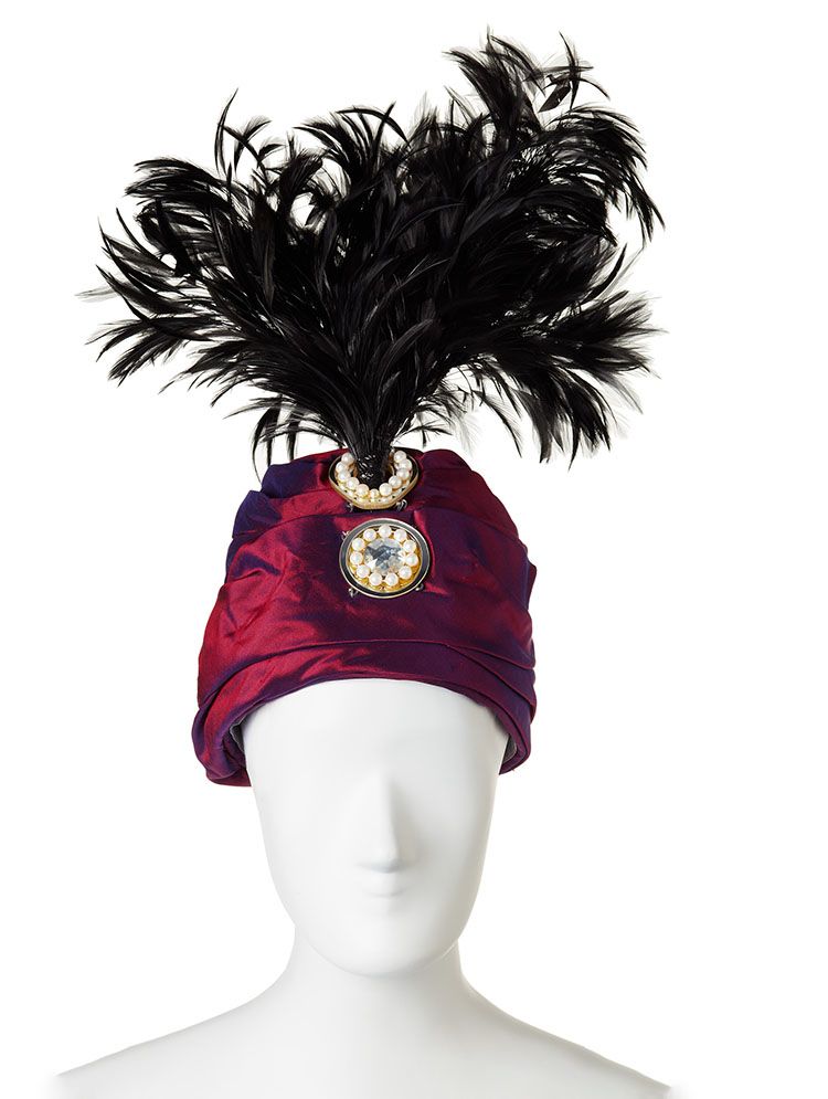Front, close-up of headdress on white mannequin. It is a turban-like hat in purple mettalic fabrics with jewels at the fron and a plume of black feathers stems from the top.