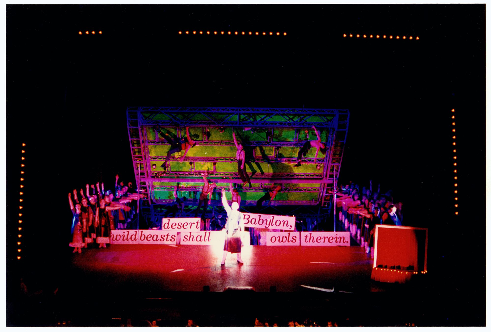 Photograph of a theatre stage with cast members in two lines on each side, as well as handing from a green backdrop, each with a fist in the air. In the foreground a single actor repeats the stance in front of signs indivudally reading: 'wild beasts', 'desert', 'shall', 'Babylon,' 'owls', and 'therein.'
