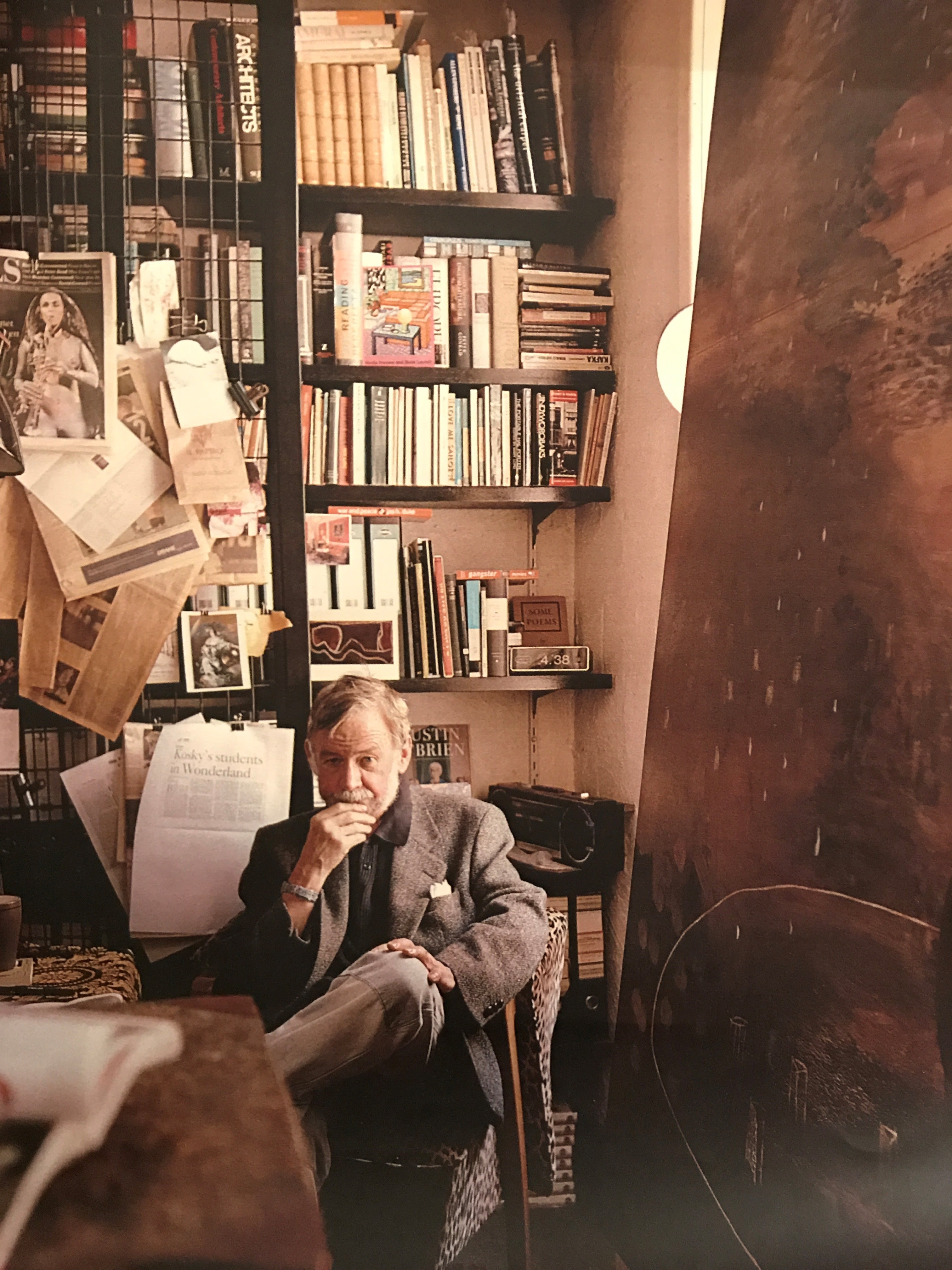 Colour potrait of man, sitting cross legged. He is surrounded by books and paper material.