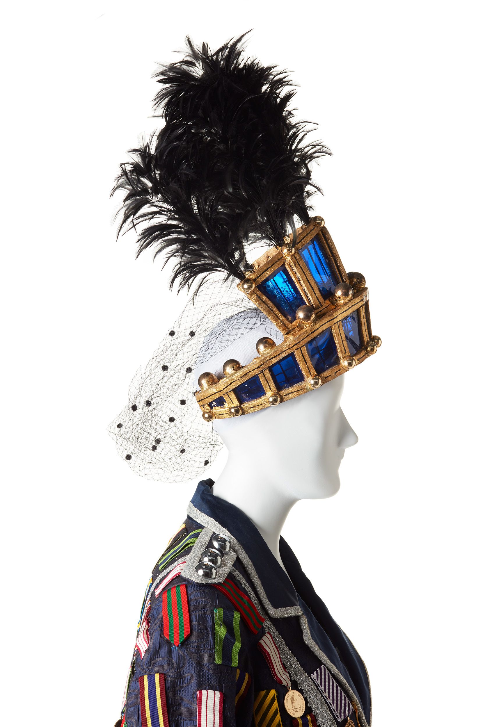 Close up shot of costume and headdress on white mannequin. It features a navy blue, floor lenght coat with medals all over. A headdress of gold and blue is topped with black feathers and features a black netting on the back..