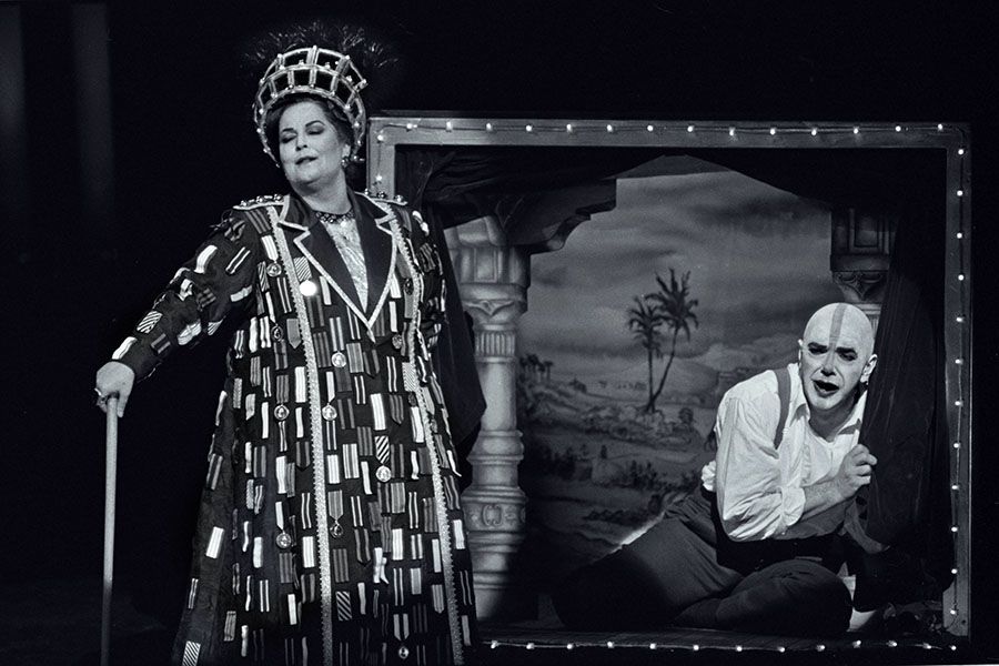 Black and white photograph of a man and woman on stage in costume. The woman is wearing a decorated coat, feathered headdress and cane. The man is sitting in a miniature theatre, his face is painted white.