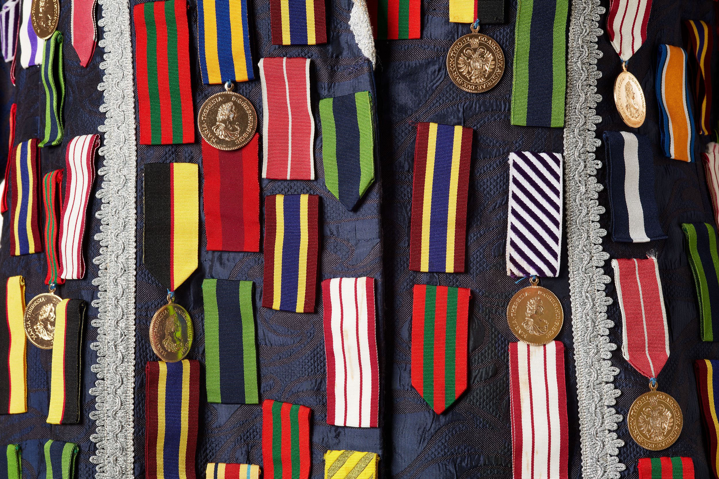 Close up of navy blue brocade material covered in coloured medals, featuring silver lace trimming.