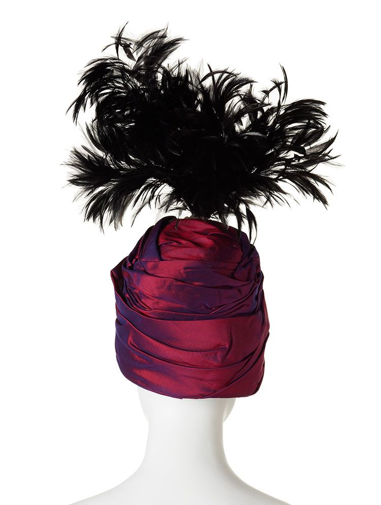 Back, close-up of headdress on white mannequin. It is a turban-like hat in purple mettalic fabrics a plume of black feathers stems from the top.