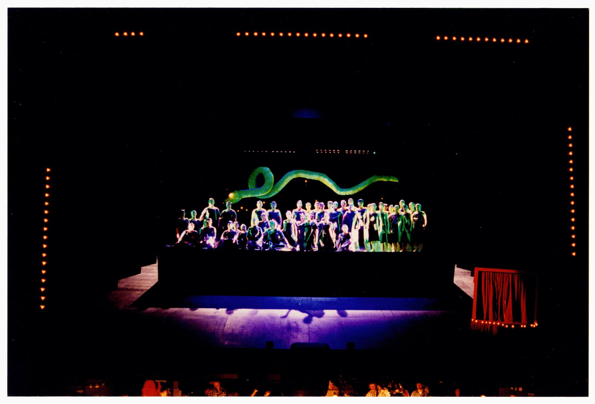 Photograph of theatre stage with cast on raised platform beneath a green snake like figure.