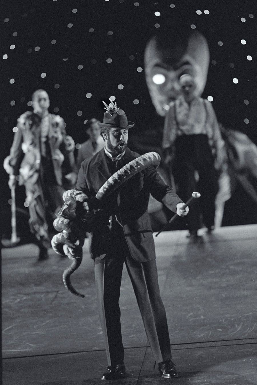 Black and white photograph of a theatre stage. A man in a suit carrys a cane and has a prop snake wrapped around his body and arms. His hat featues a prop insect. Other actors appear blurred in the background.