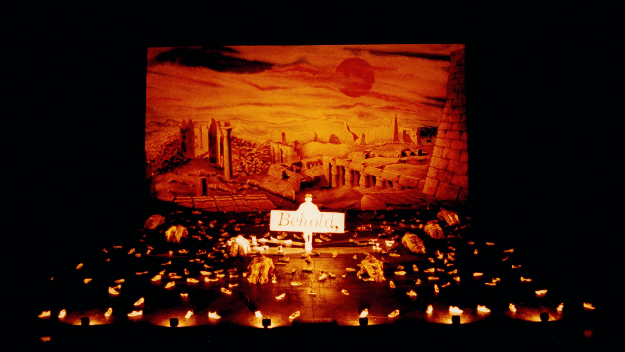 Photograph of theatrical set. A red stone building landscape features in the background. On stage a man in white hold a sign reading 'Behold,'. Actors in white, and single white shoes lay on the entirety of the stage.