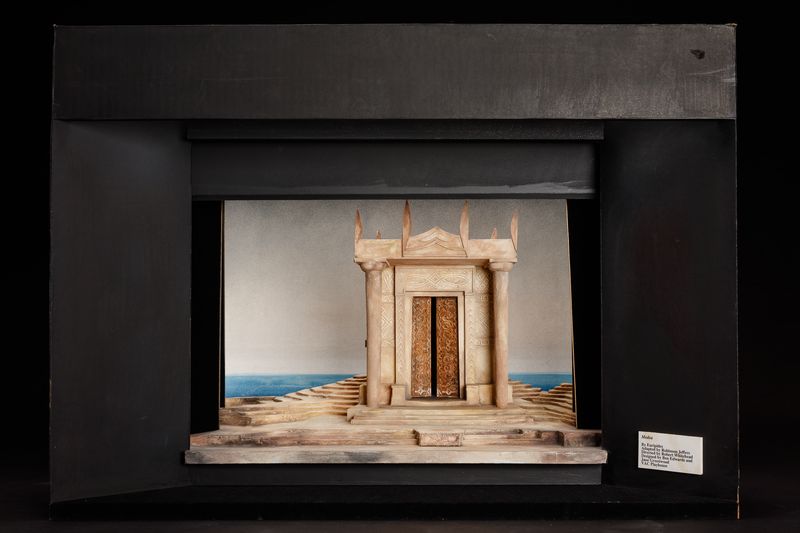 A small Greek temple in a stony landscape by the sea, with its wooden doors partly open.